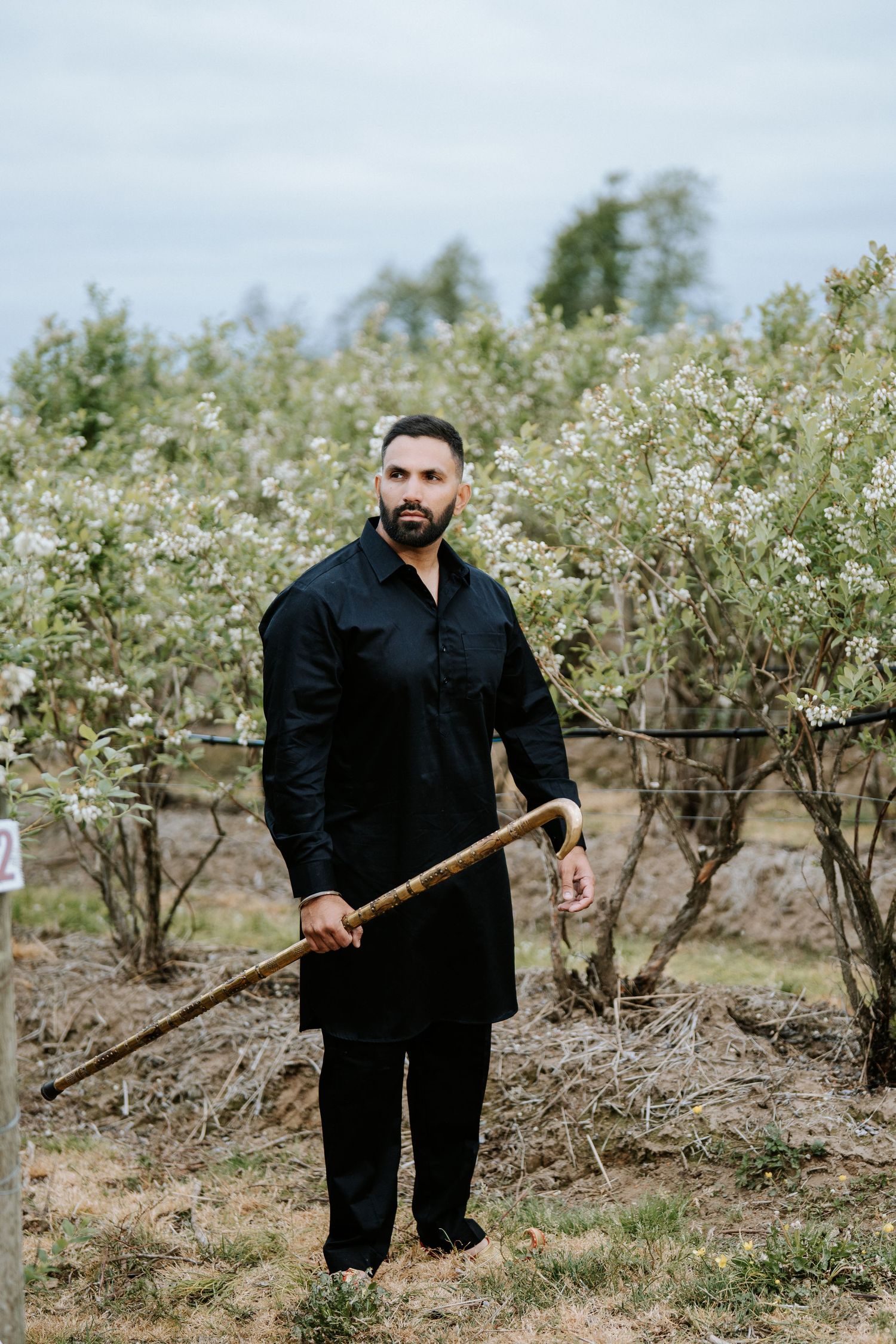 Man in black kurta holding a wooden staff among flowering trees in an orchard.