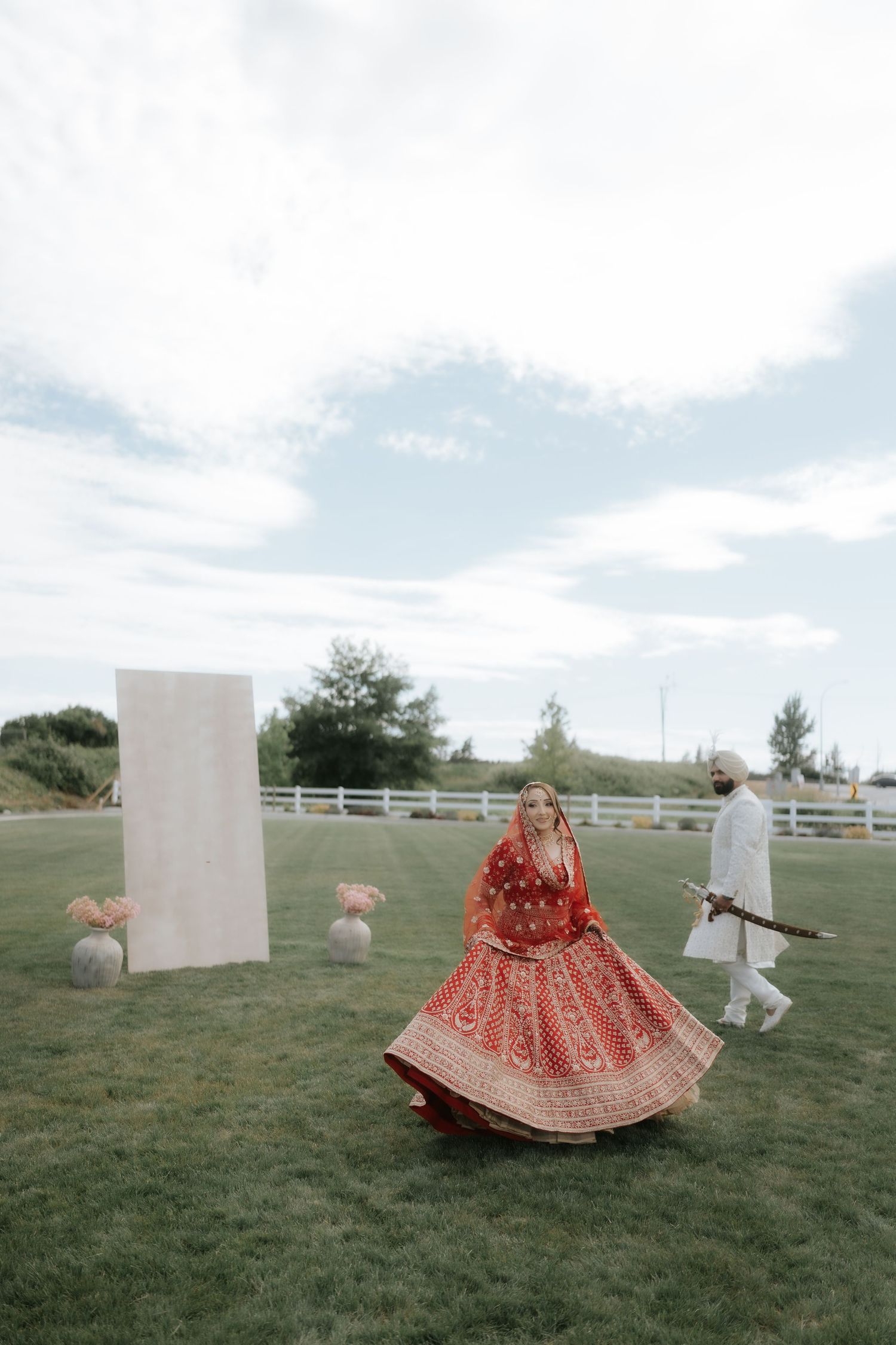 Bride in red lehenga twirling on green lawn at outdoor Indian wedding ceremony with groom in white sherwani.