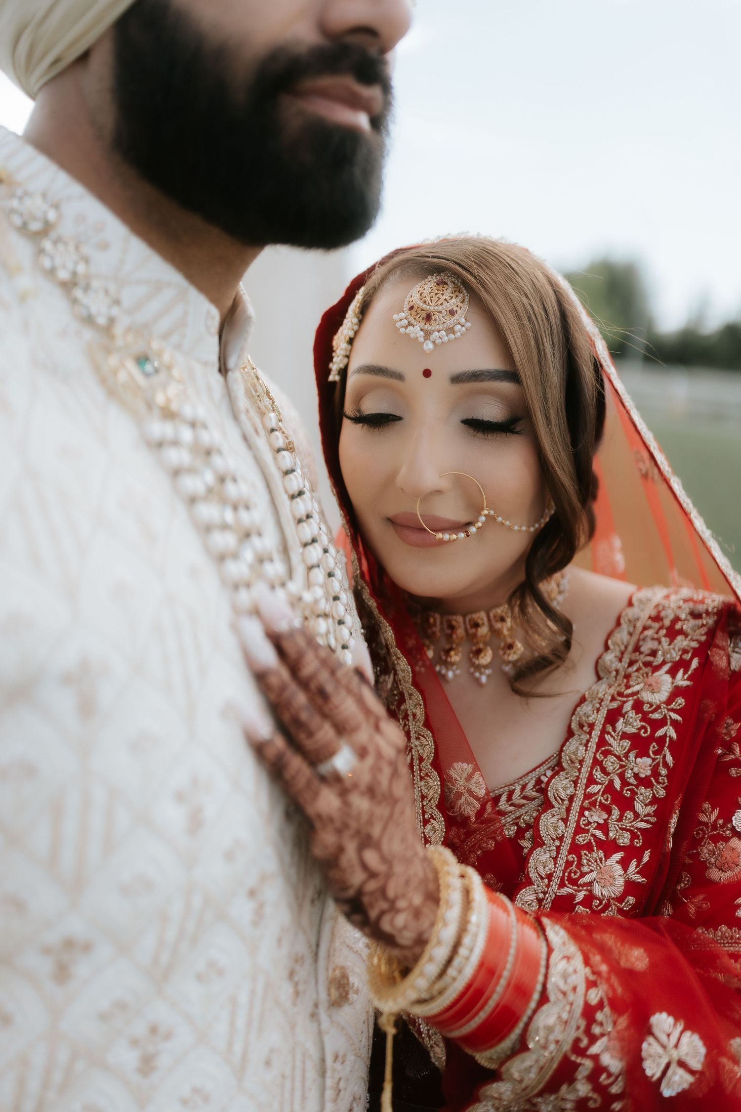 Indian bride in red bridal lehenga with gold jewelry and maang tikka leans against groom in white sherwani.