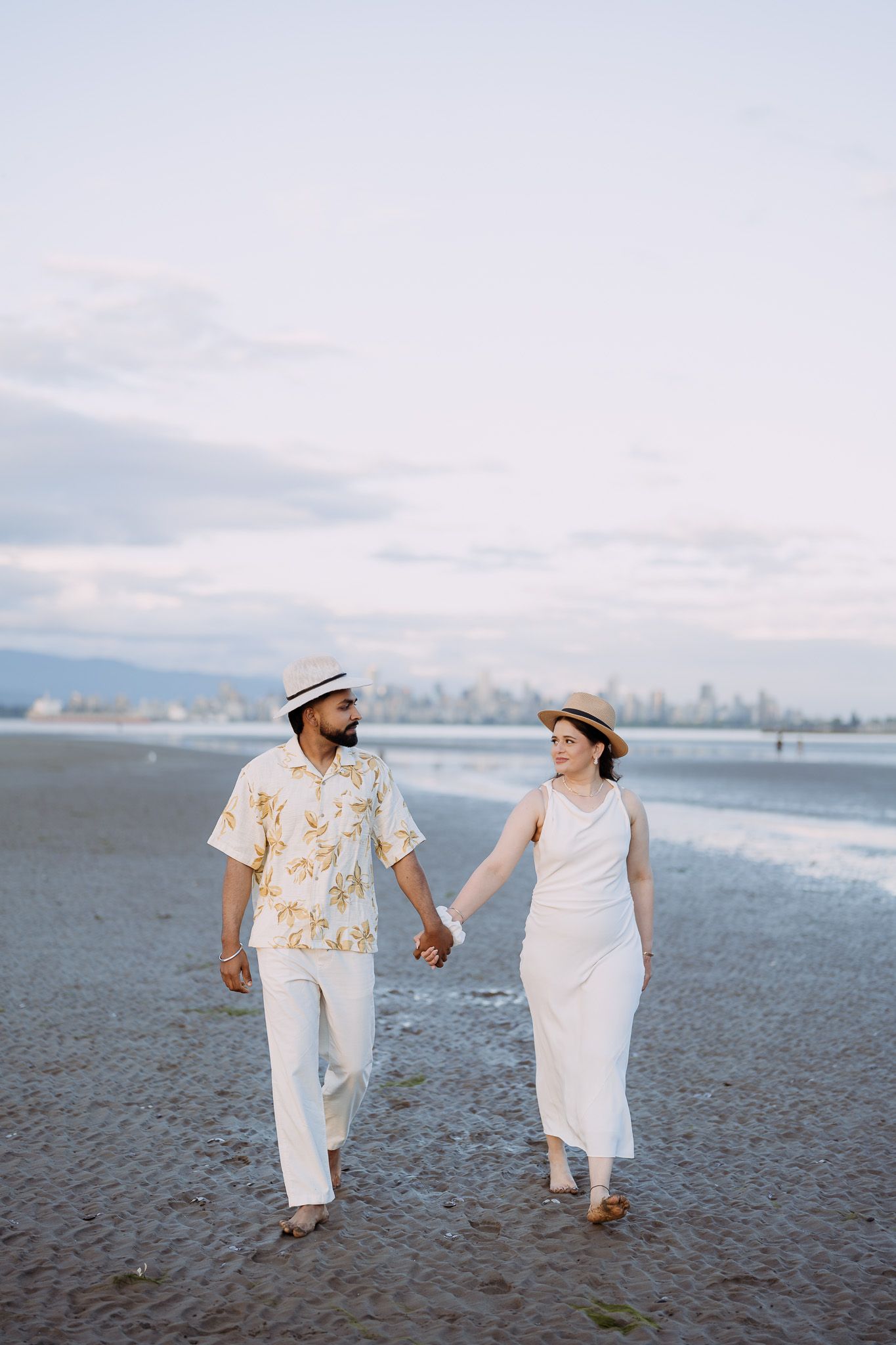 Happy couple in white outfits and straw hats holding hands while walking on a beach at dusk.