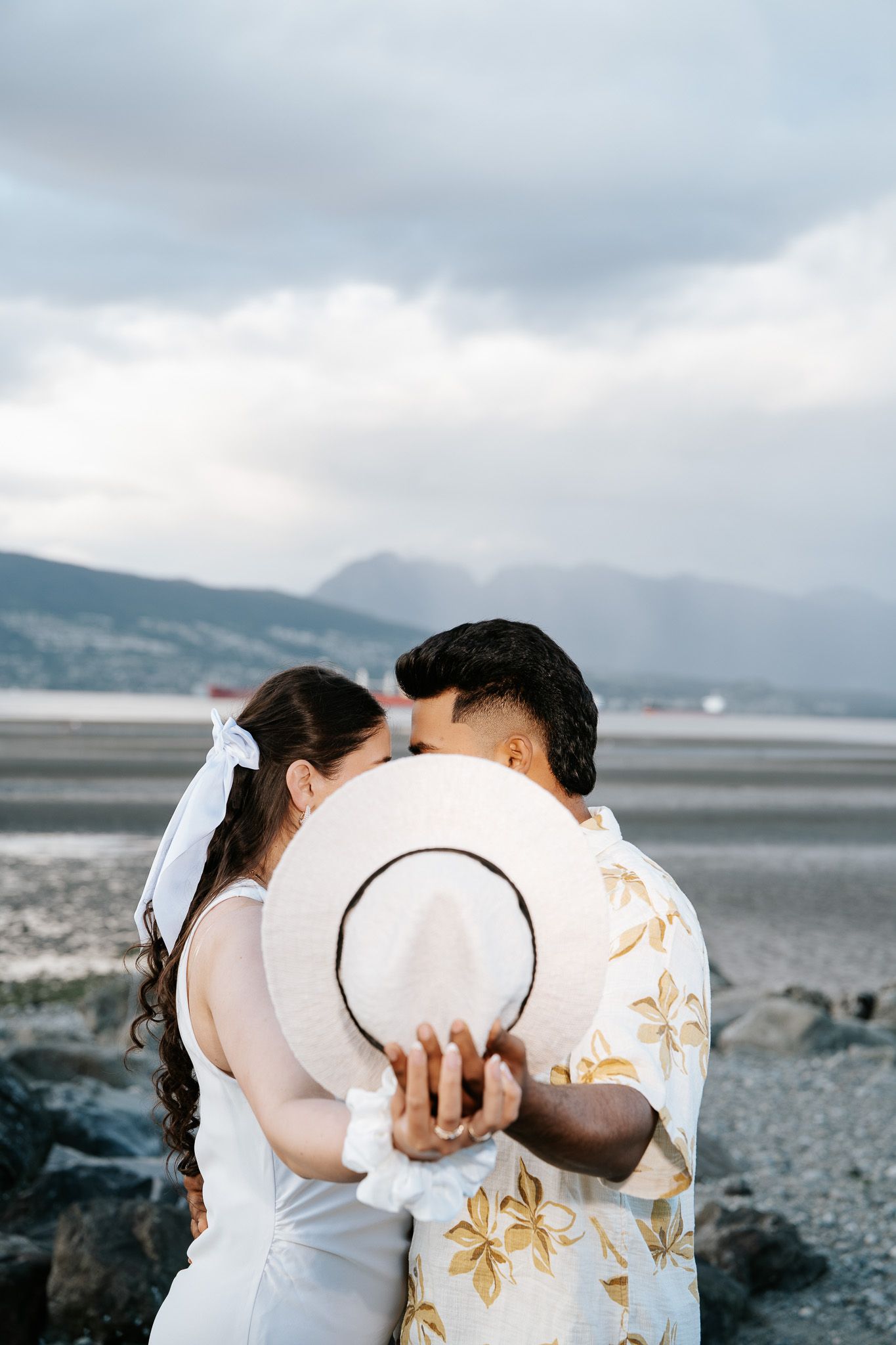 Couple kissing behind a white hat on a rocky beach with mountains and cloudy sky in the background.