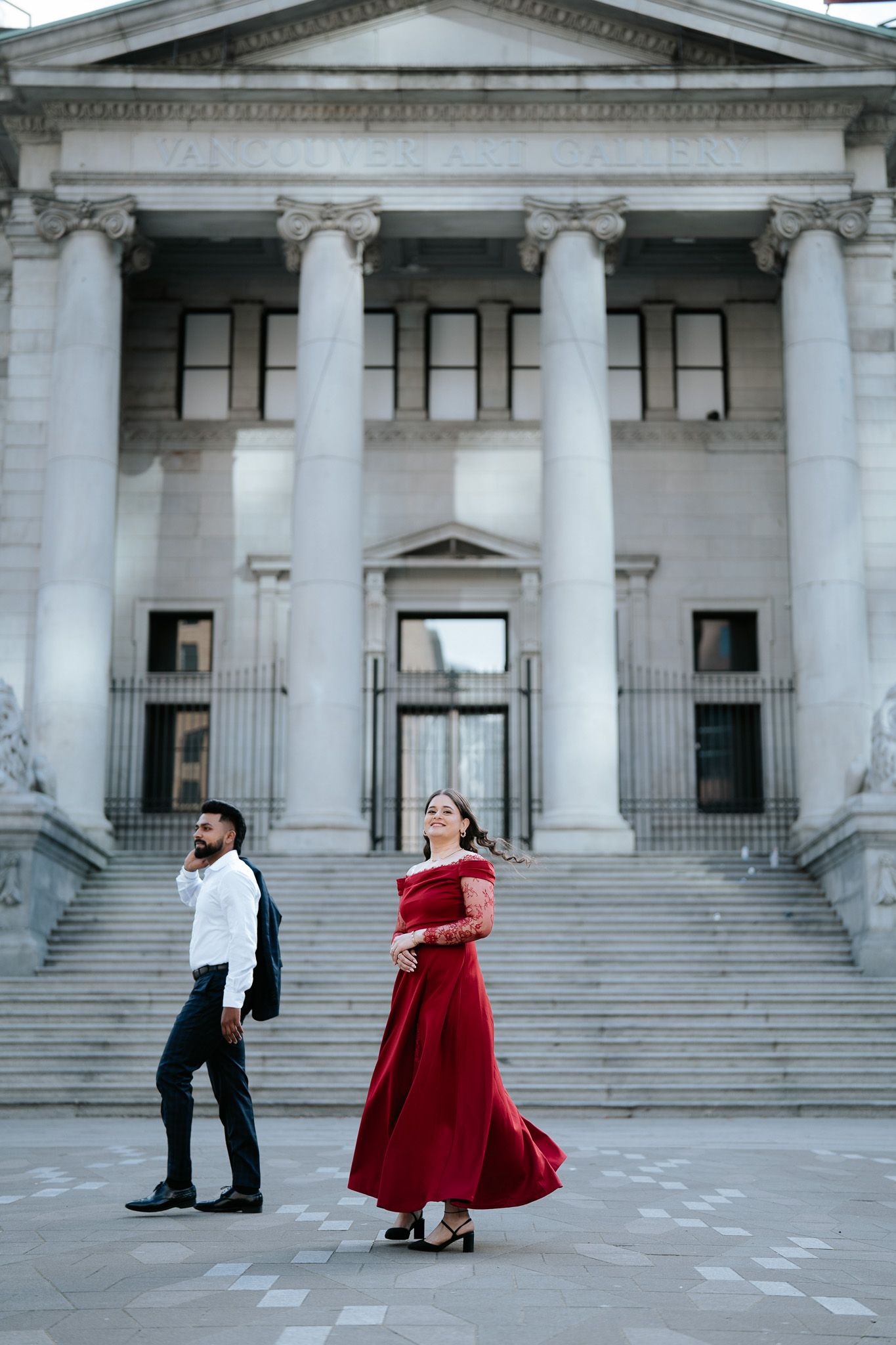 Couple in formal wear walking in front of a grand neoclassical building with tall columns, woman in red gown.