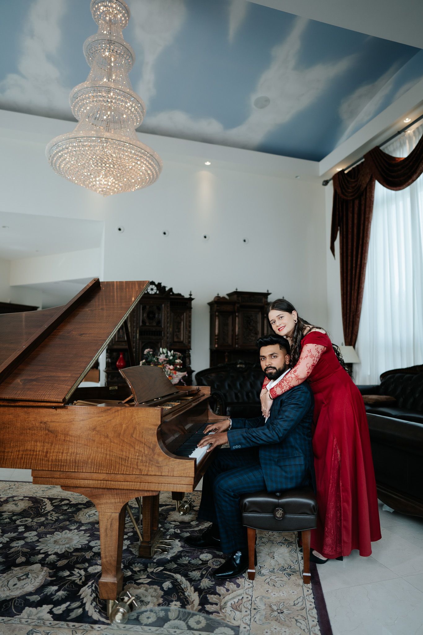 Elegant couple posing at a grand piano beneath a crystal chandelier with cloud-painted ceiling.