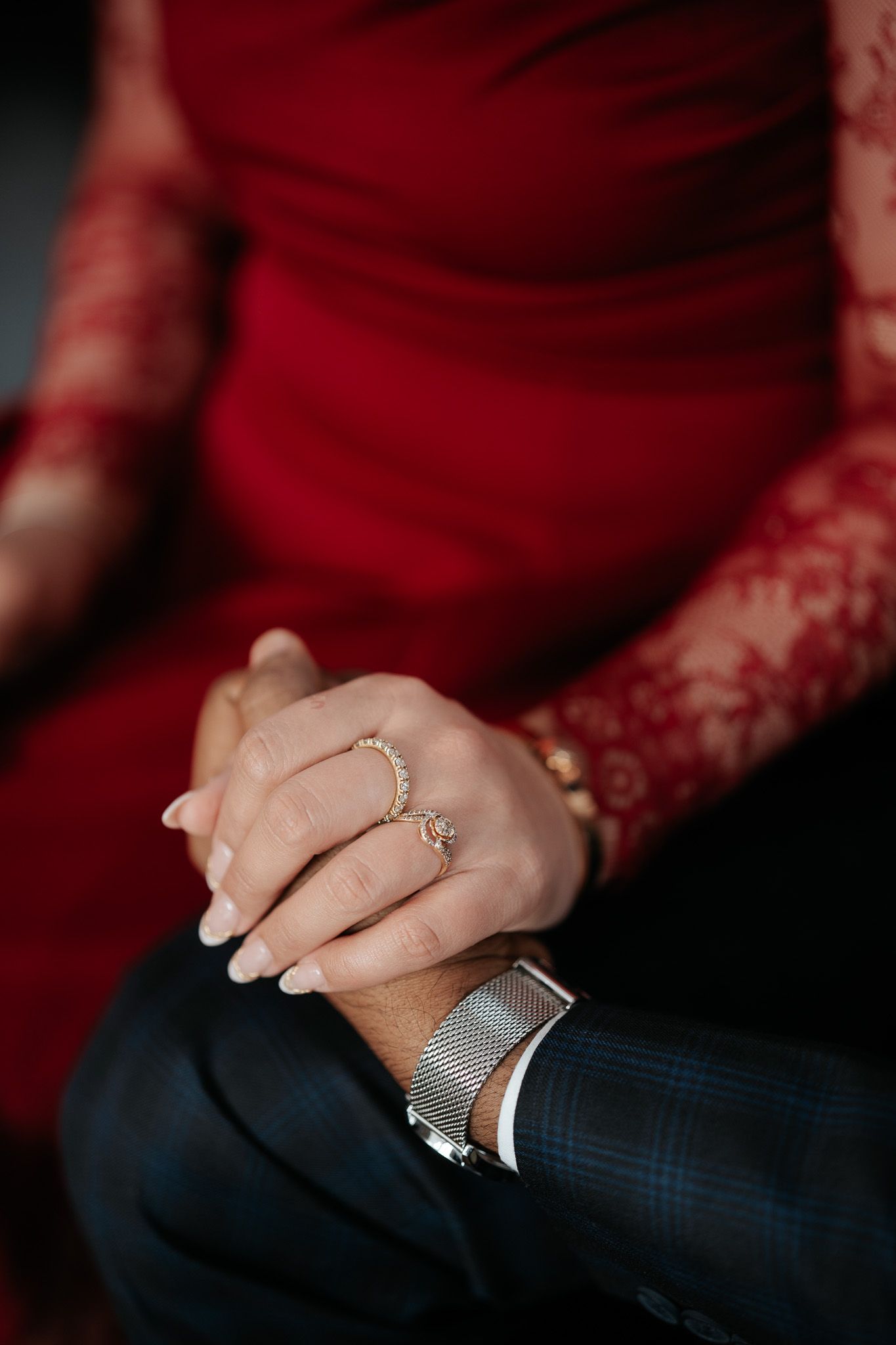 Close-up of couple holding hands, woman wearing gold ring and bracelet in red lace dress, man in plaid suit with silver watch.