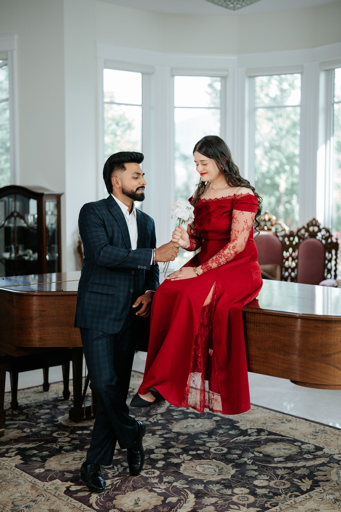 A bearded man in a plaid suit offers a flower to a woman in a red lace gown seated on a grand piano.