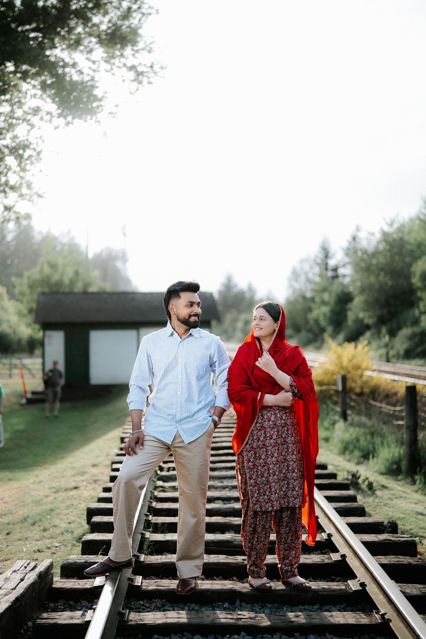 Couple standing on railway tracks outdoors, man in light blue shirt and woman in red traditional attire.