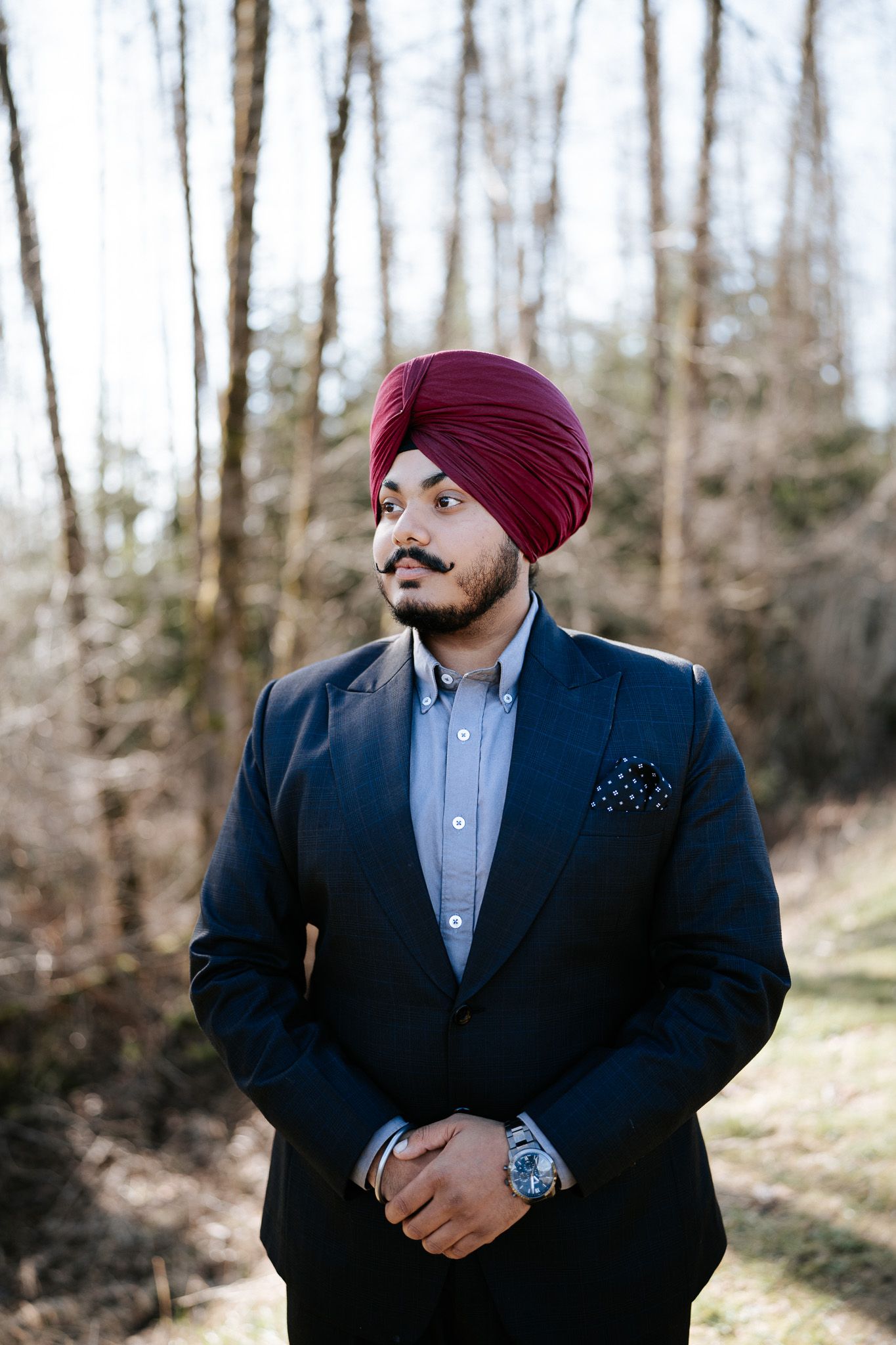 A person in a navy suit and burgundy turban stands in a wooded setting with a polished, professional appearance.