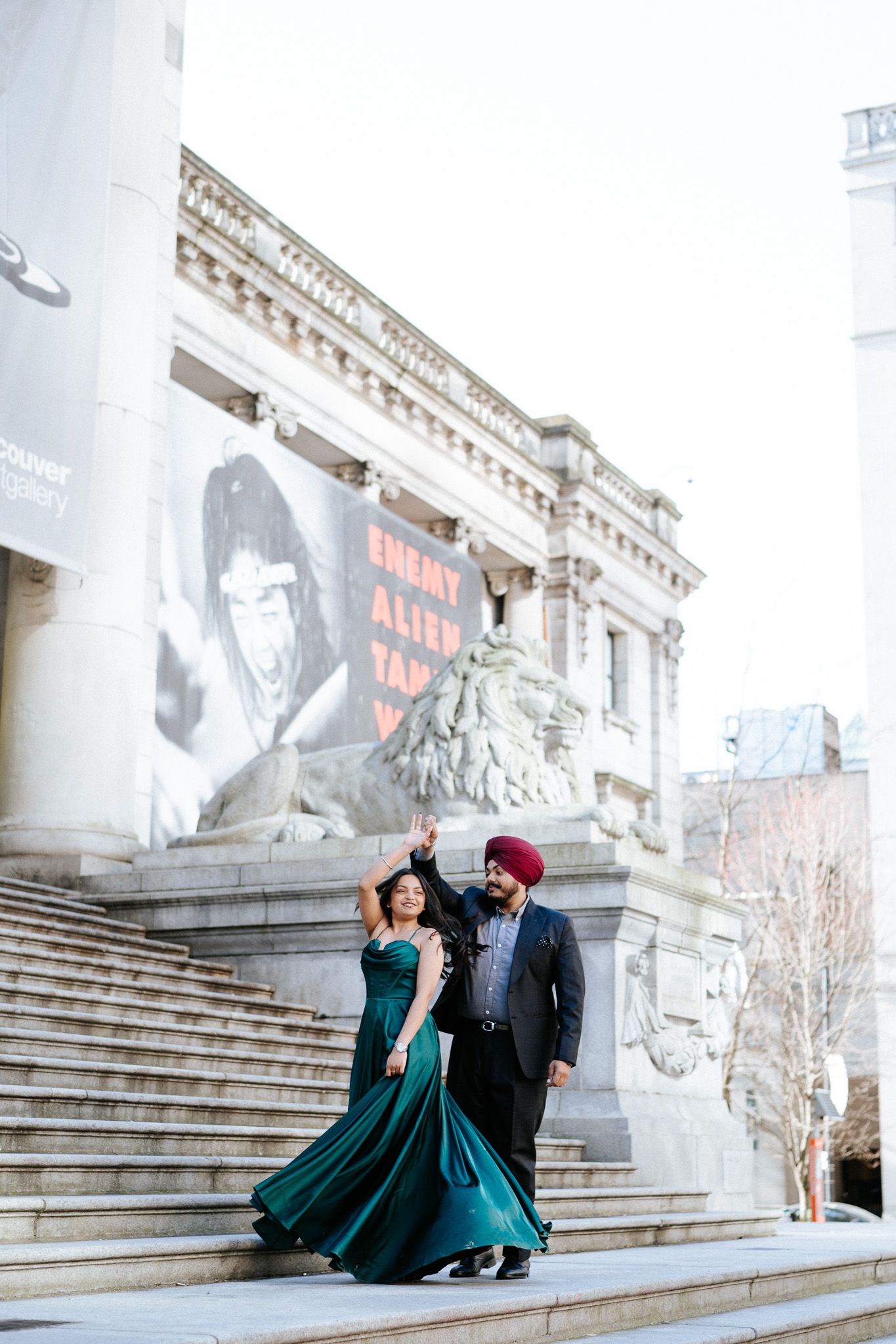 Couple in formal attire poses on grand stone steps, with person wearing elegant emerald green gown.