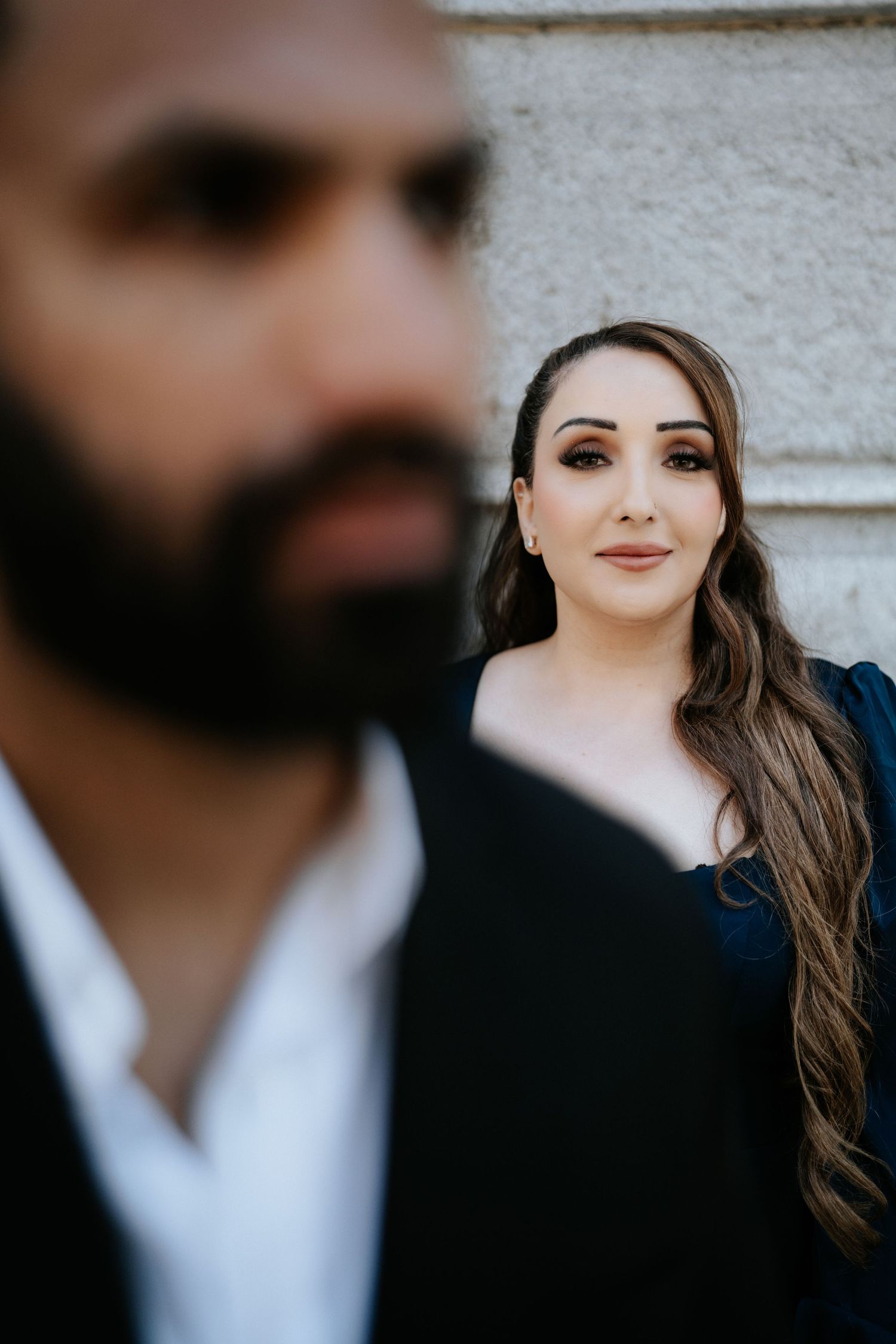 A woman with long hair and elegant makeup smiles softly behind a bearded man in a black suit during a couples portrait.