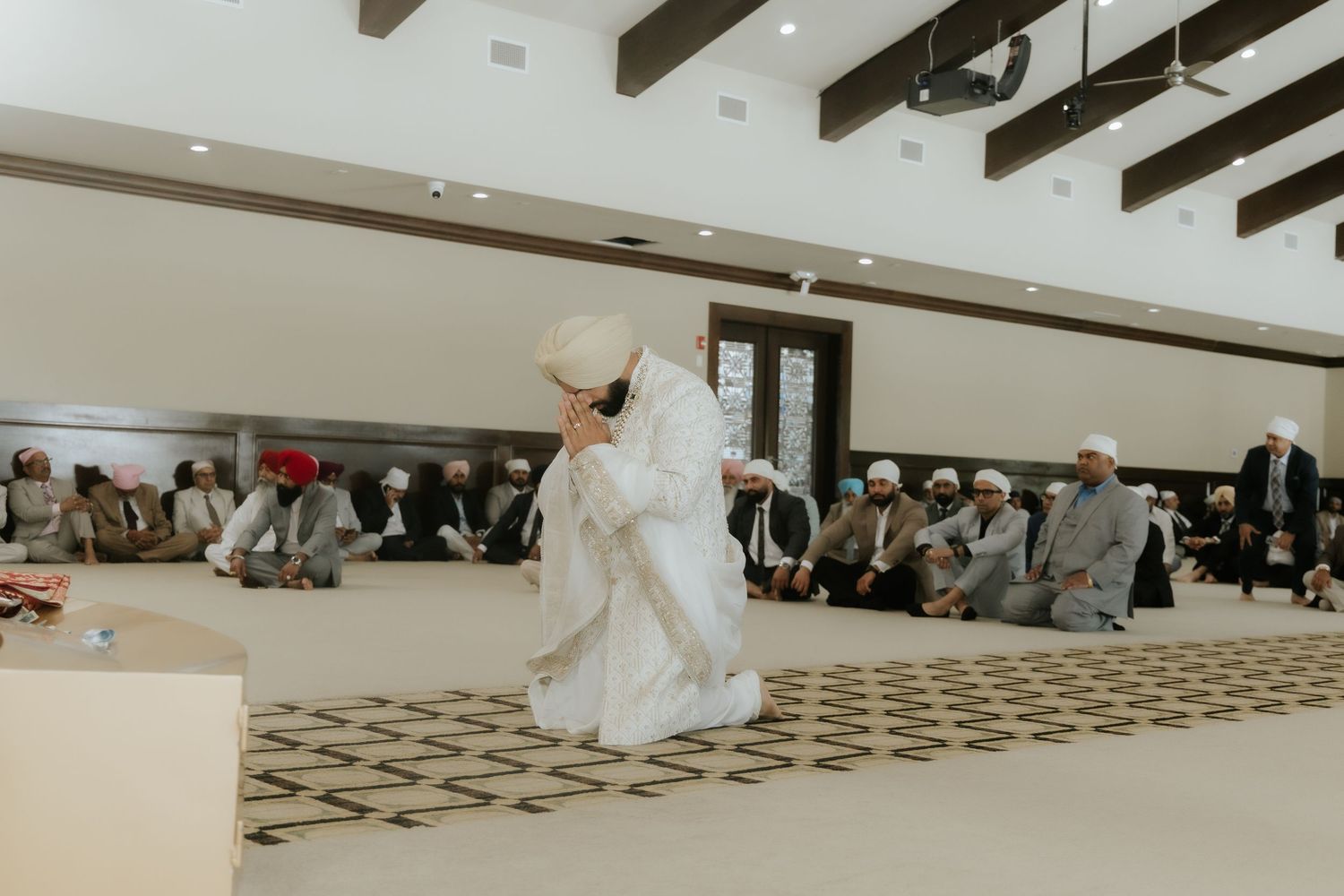 A Sikh ceremony in a modern gurdwara with worshippers seated on the floor and a person in white attire performing a ritual.