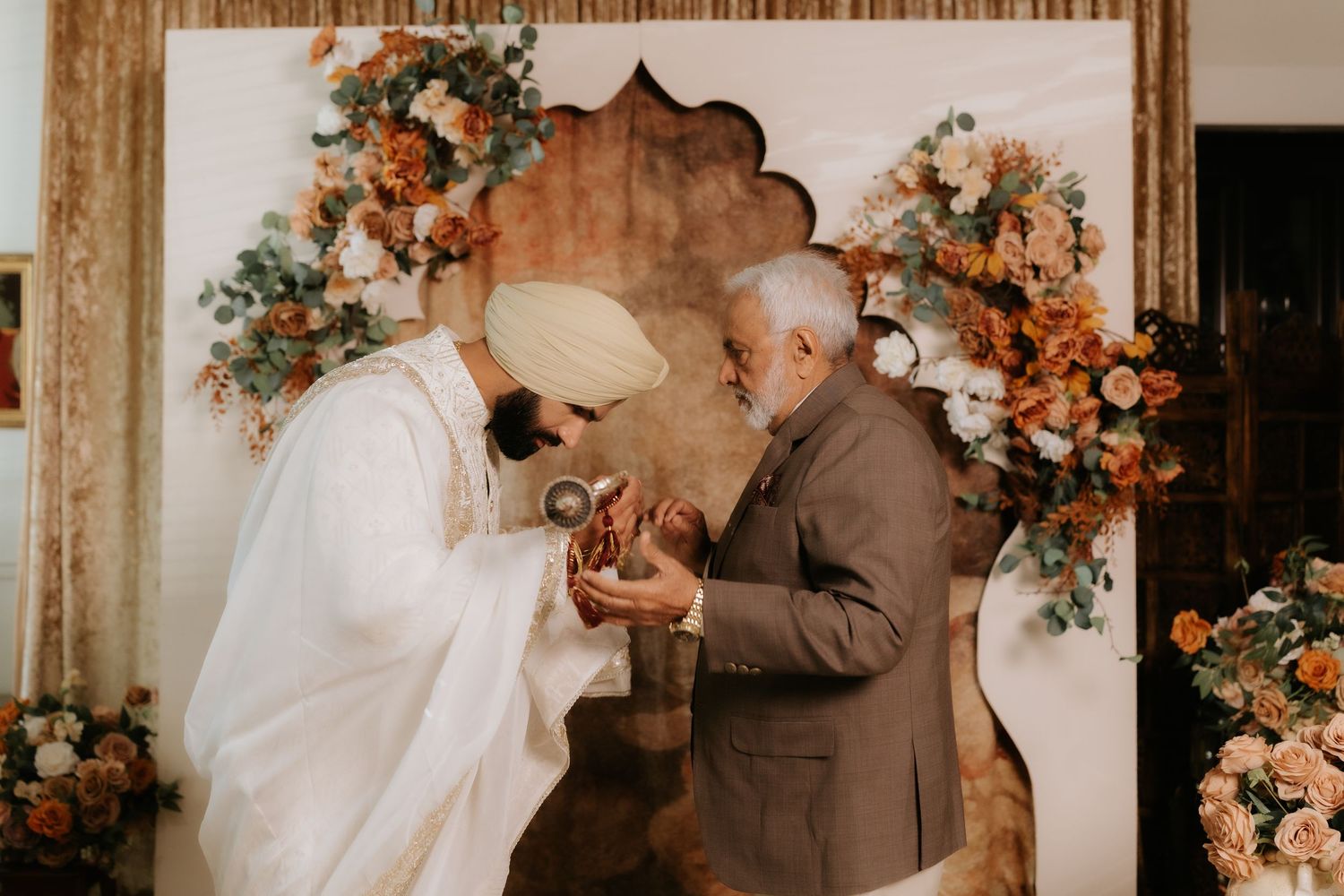 Sikh groom in white turban exchanges rings with elder at floral wedding ceremony backdrop with orange blooms.