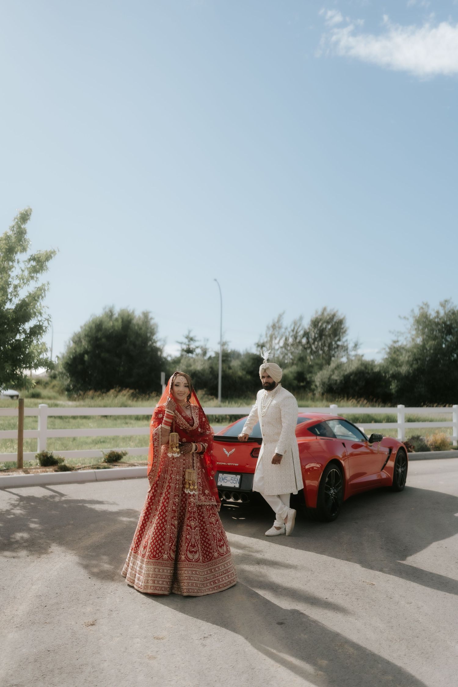 Indian bride in red lehenga and groom in white sherwani pose beside a red Corvette sports car outdoors.