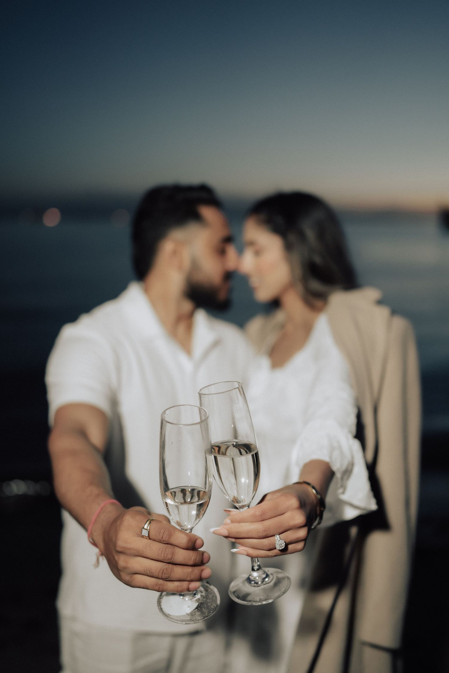 Two champagne glasses clink together against a blurred ocean sunset backdrop in a romantic seaside moment.