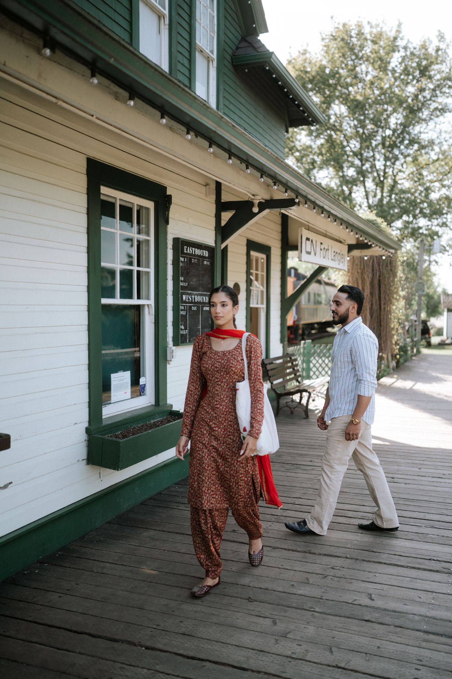 Two people walk along a wooden porch of a historic building with white walls and green trim.