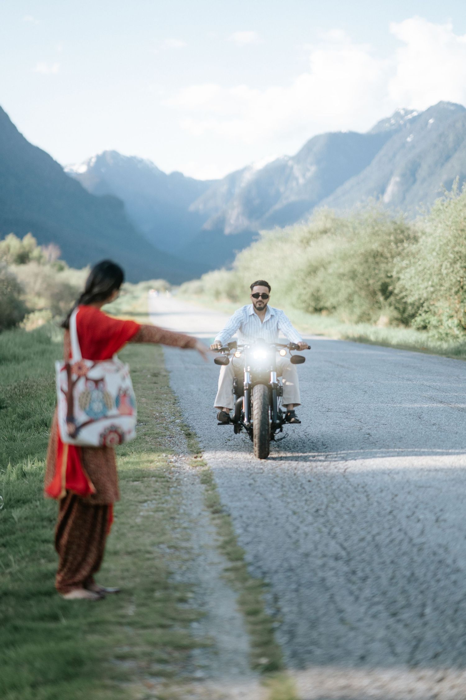 A motorcyclist rides along a scenic mountain road while a person in red stands by the roadside watching.