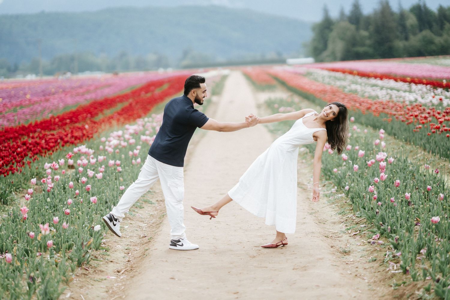 A couple walks through vibrant red and pink tulip fields with mountains in the background on a sunny day.