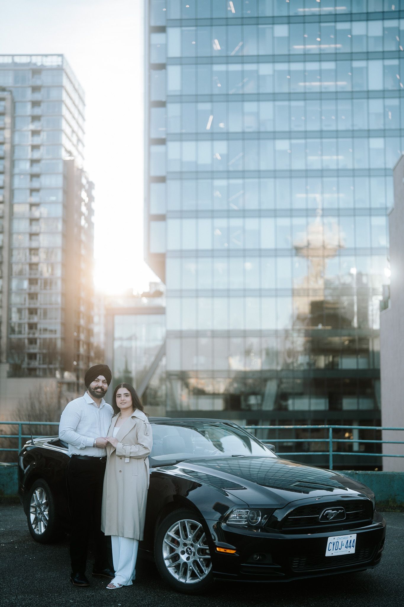 A black luxury car parked in front of a modern glass building at sunset with reflective surfaces.