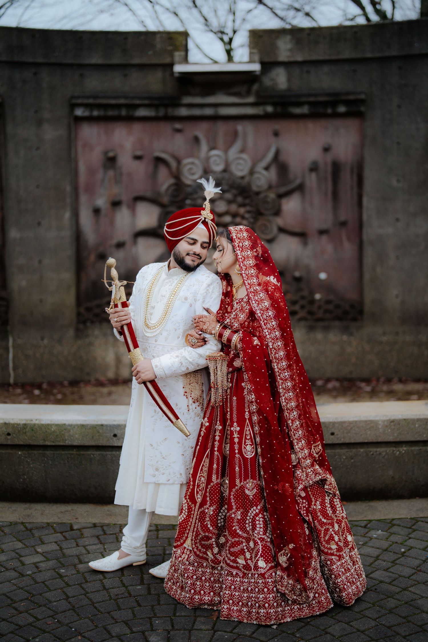 Traditional Indian wedding couple in white and red attire pose together on cobblestone ground.