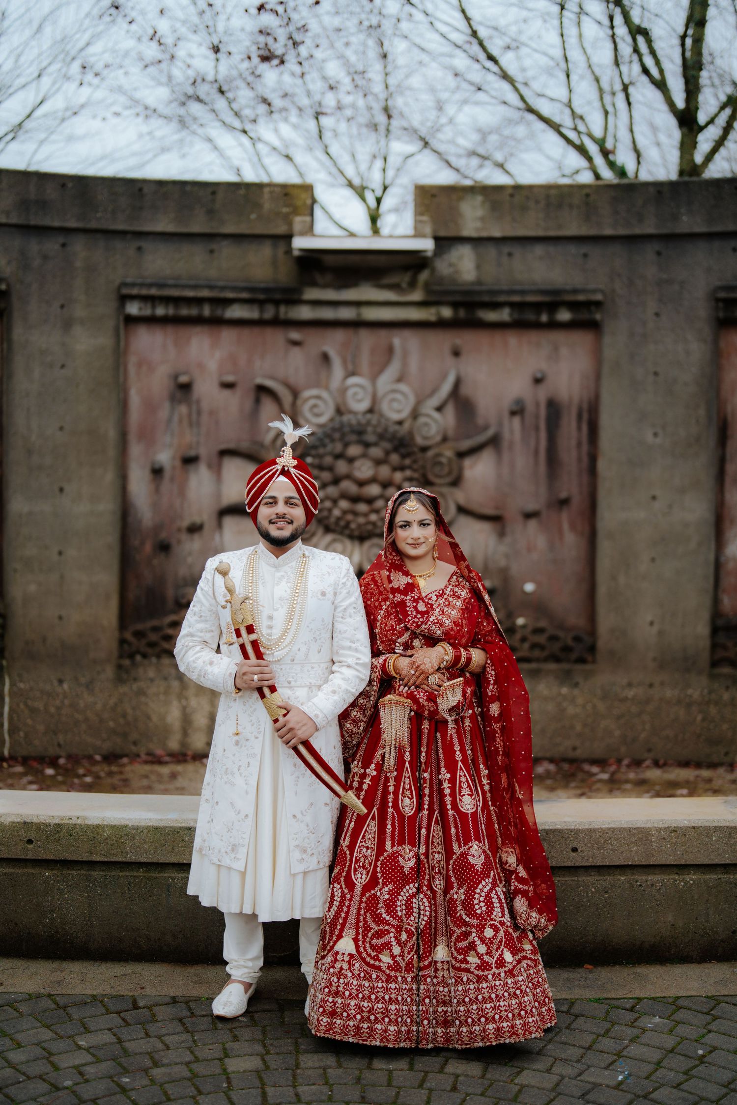 Wedding couple in traditional South Asian attire posing against stone wall backdrop.