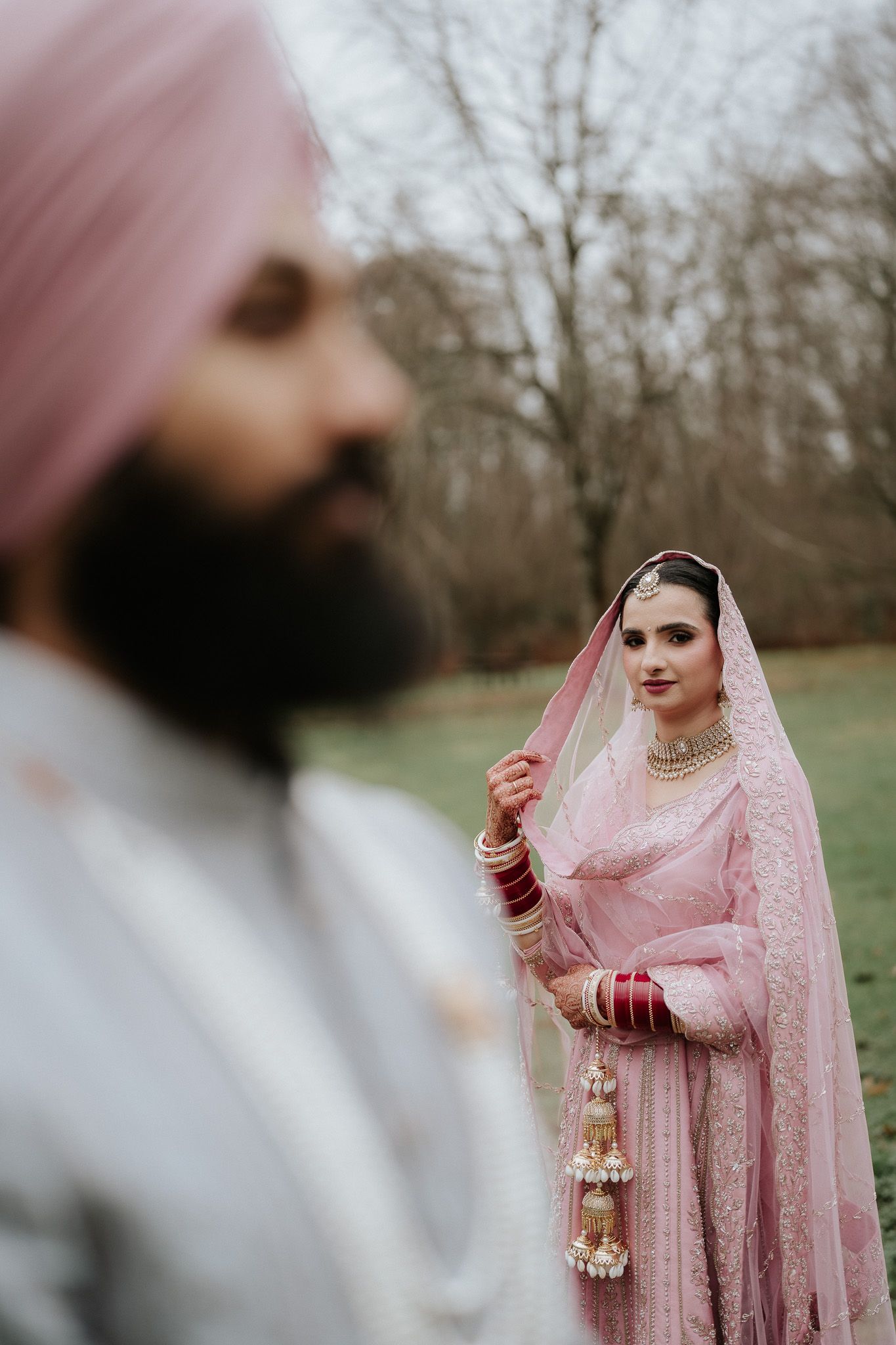 A couple in traditional South Asian wedding attire poses together in an outdoor setting with trees in the background.
