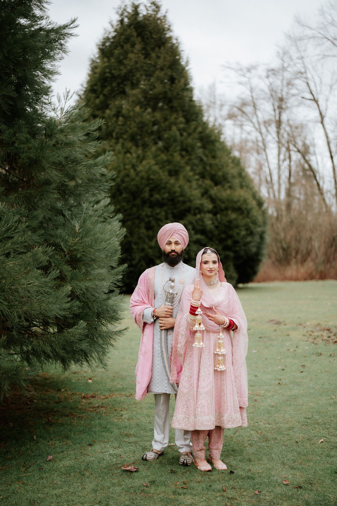 A couple in traditional pink and white South Asian wedding attire poses together in a garden setting with evergreen trees.