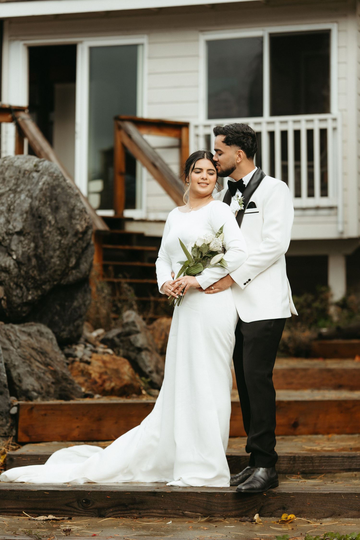 A romantic wedding couple shares intimate moments on rustic wooden steps outside a white farmhouse.