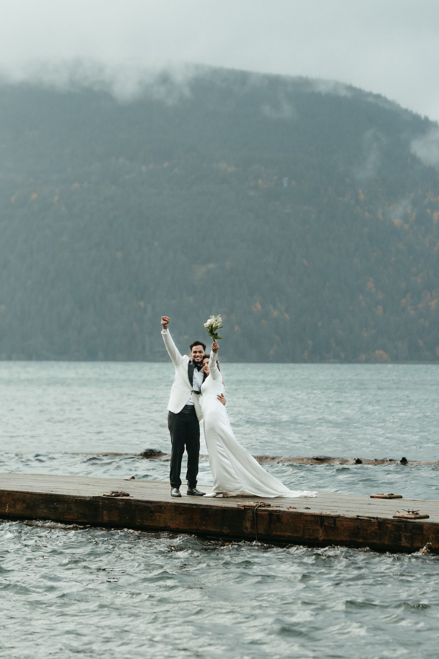 Couple poses on a wooden dock surrounded by misty mountains and lake waters on their wedding day.