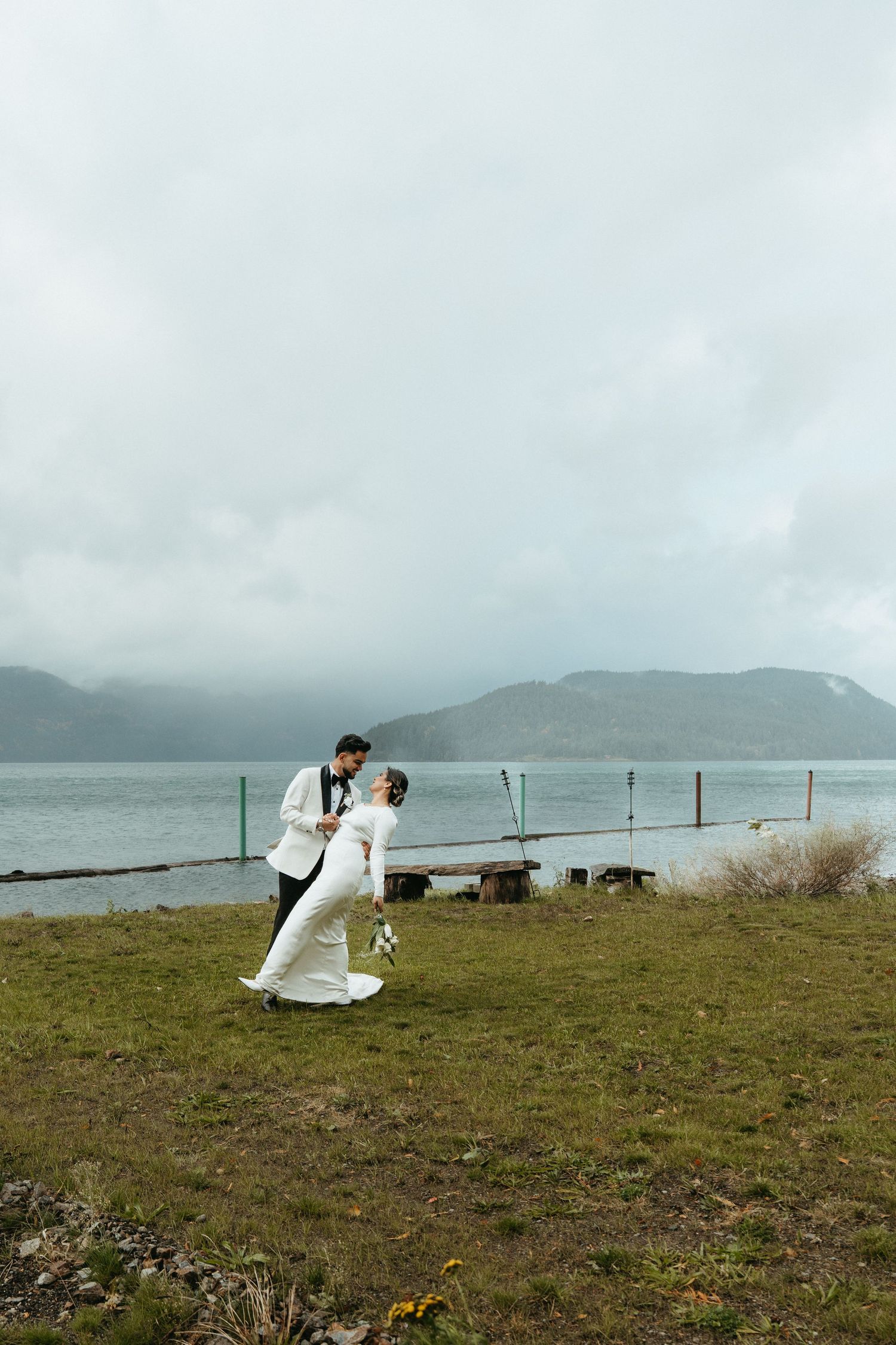 A couple in wedding attire shares a romantic moment on a scenic coastal hillside with mountains and water in the background.