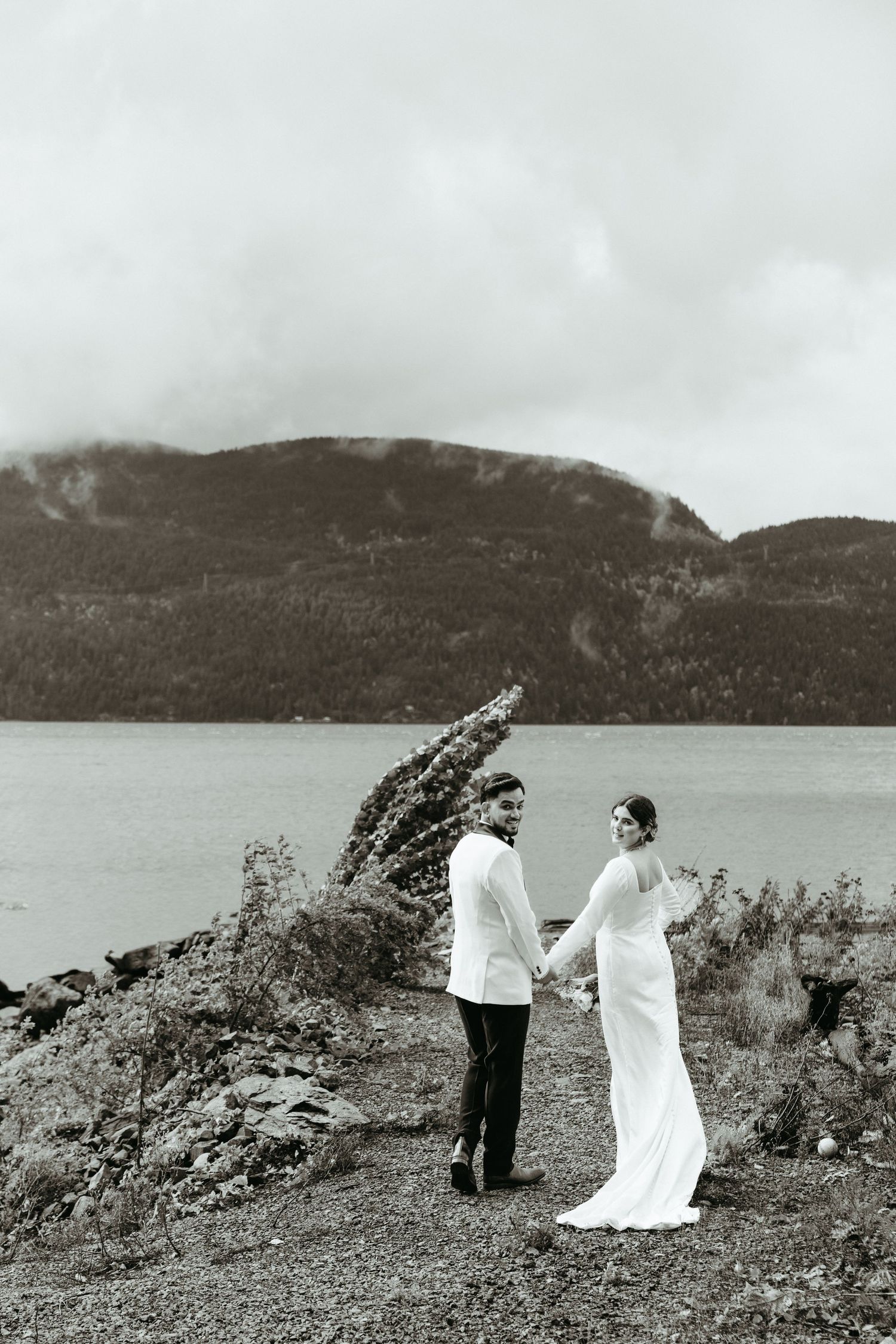 A couple in wedding attire stands on a rocky hillside overlooking a scenic lake and mountain landscape.