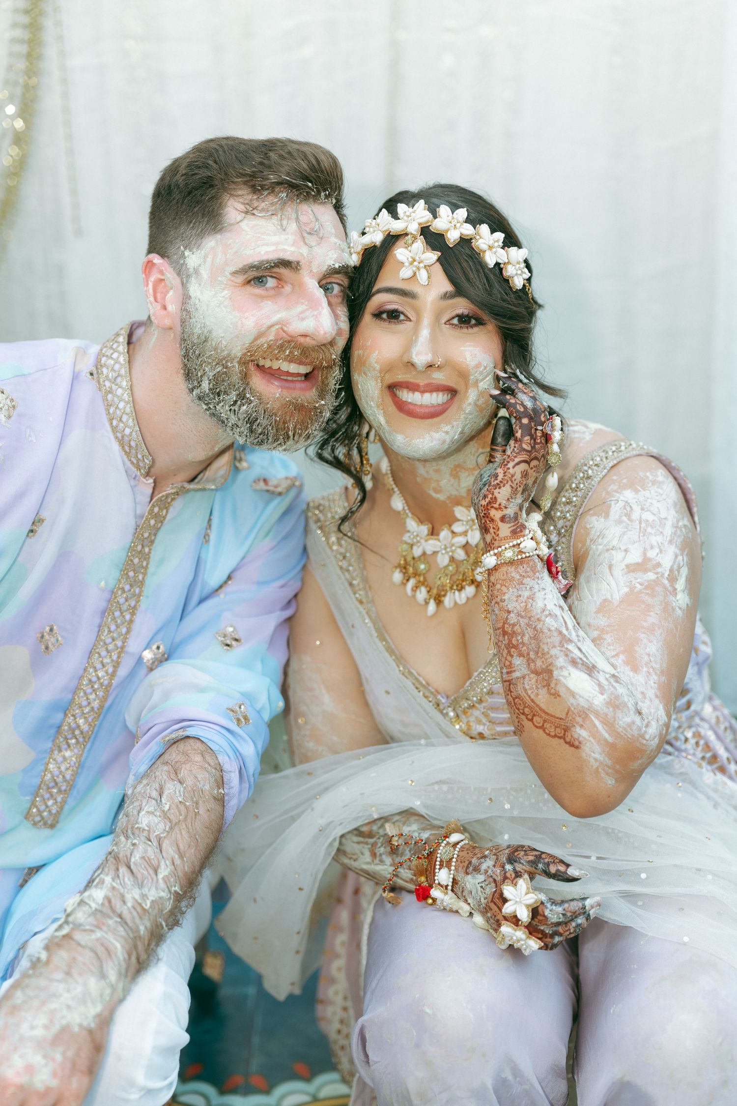 Couple covered in haldi paste smiling during Indian wedding ceremony, bride in white lehenga with floral hair accessories.