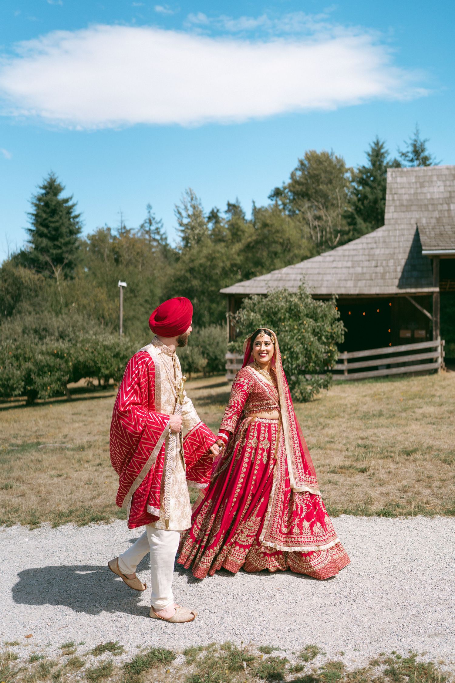 Indian bride and groom in red traditional wedding attire walking outdoors on a sunny day near a rustic barn.
