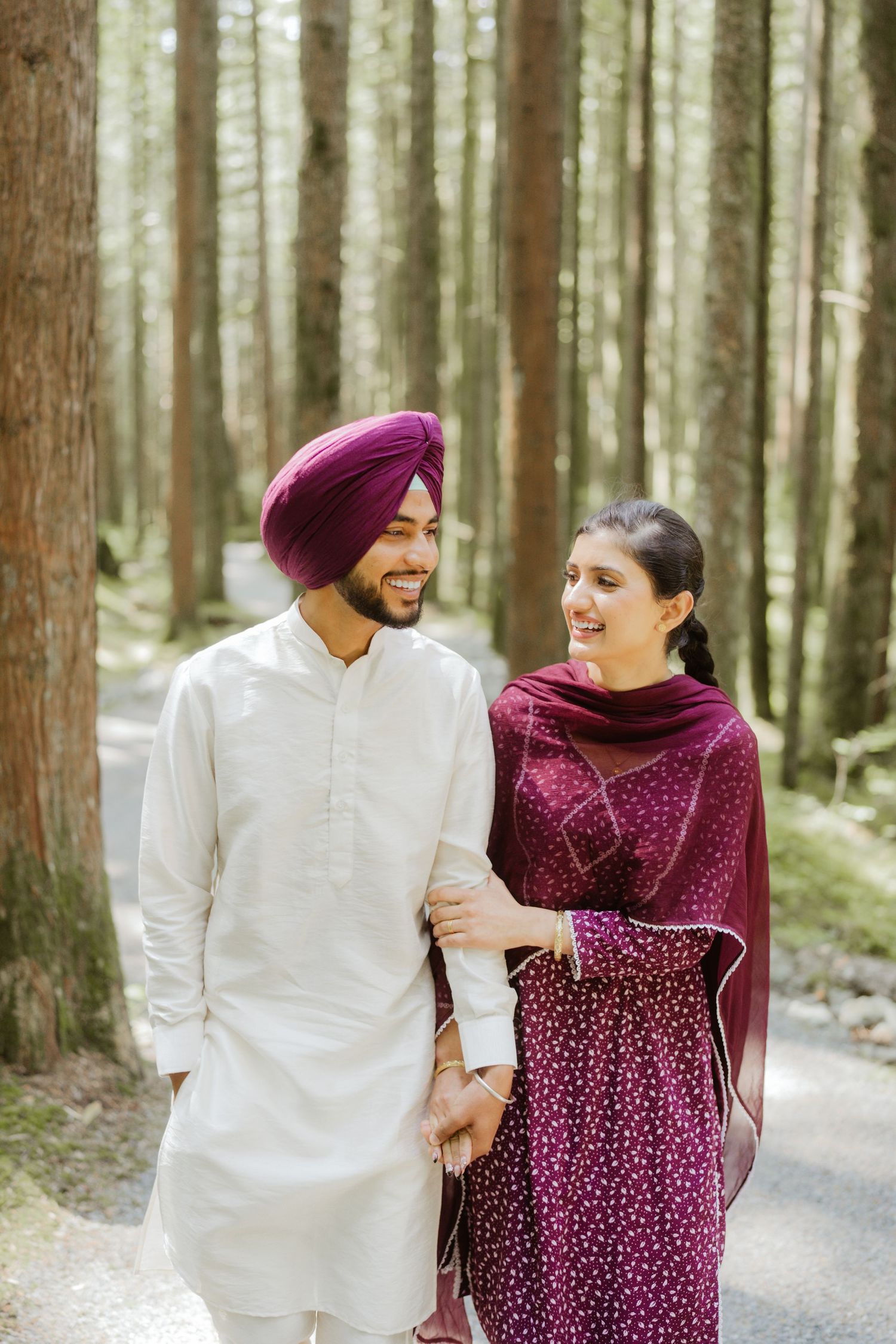 A Sikh couple in traditional attire walks hand in hand through a forest, smiling at each other.