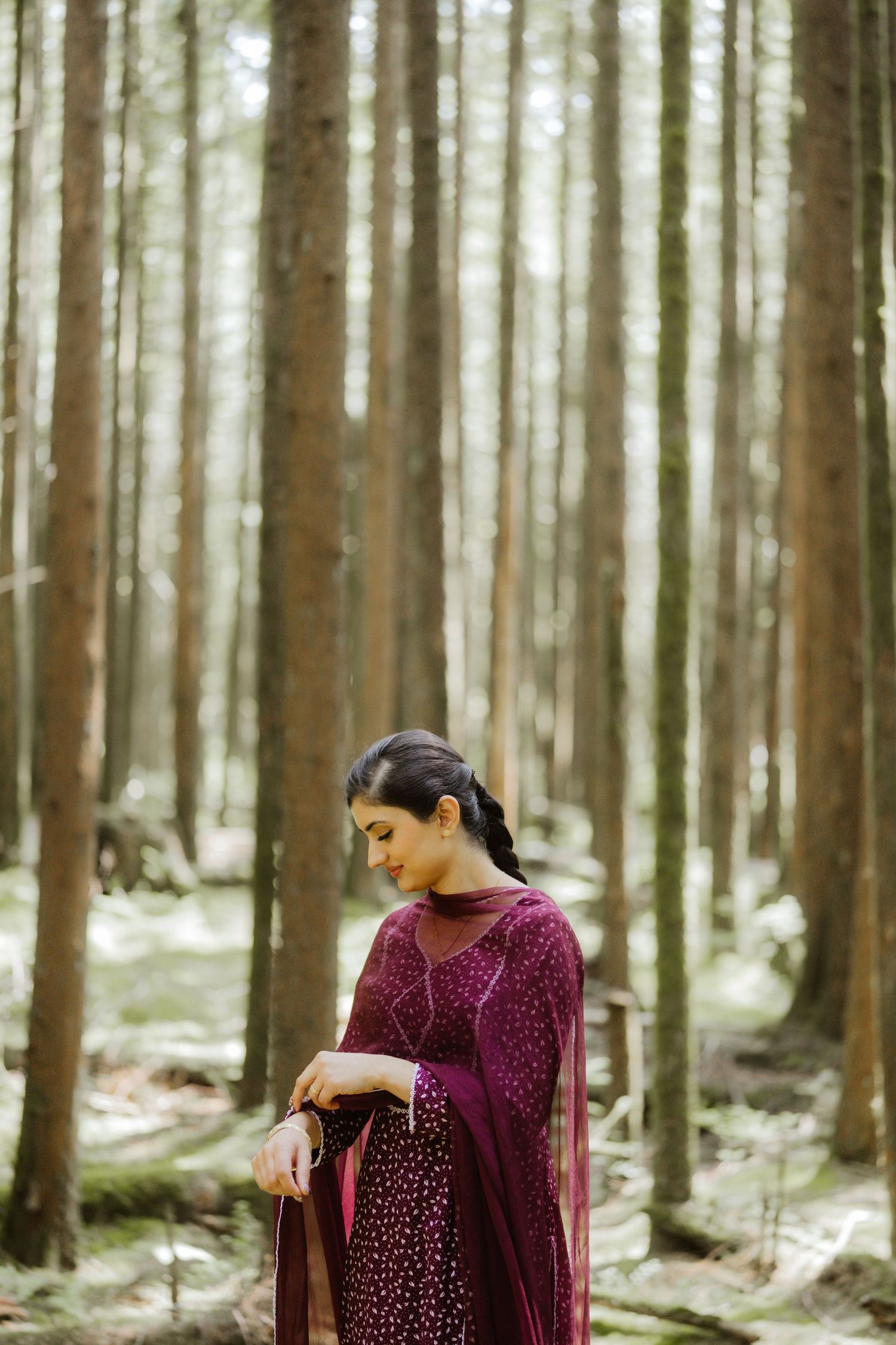 South Asian woman in burgundy embroidered salwar suit standing thoughtfully among tall trees in a forest.