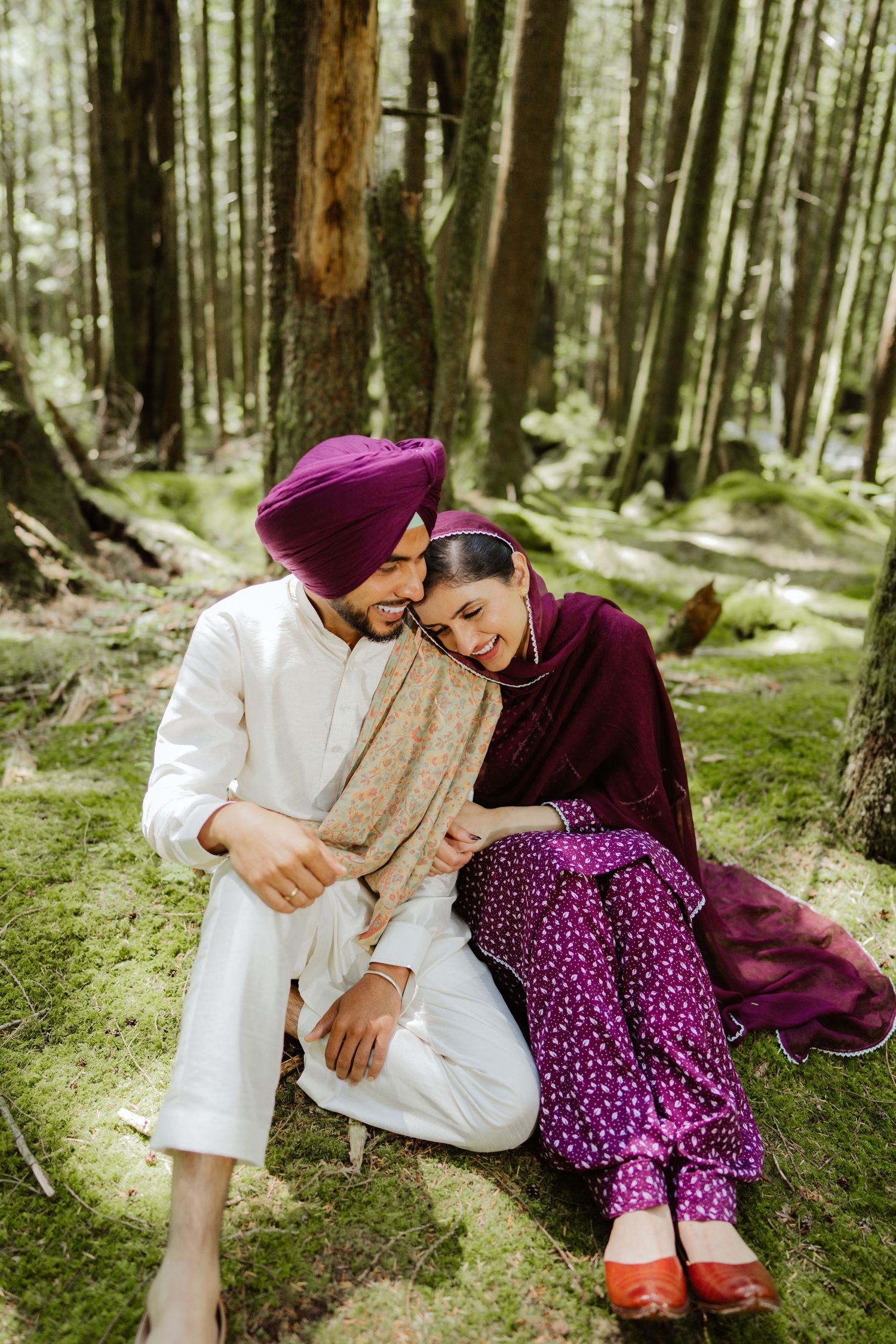 Sikh couple sitting on mossy forest ground, man in white kurta and purple turban, woman in burgundy salwar kameez.