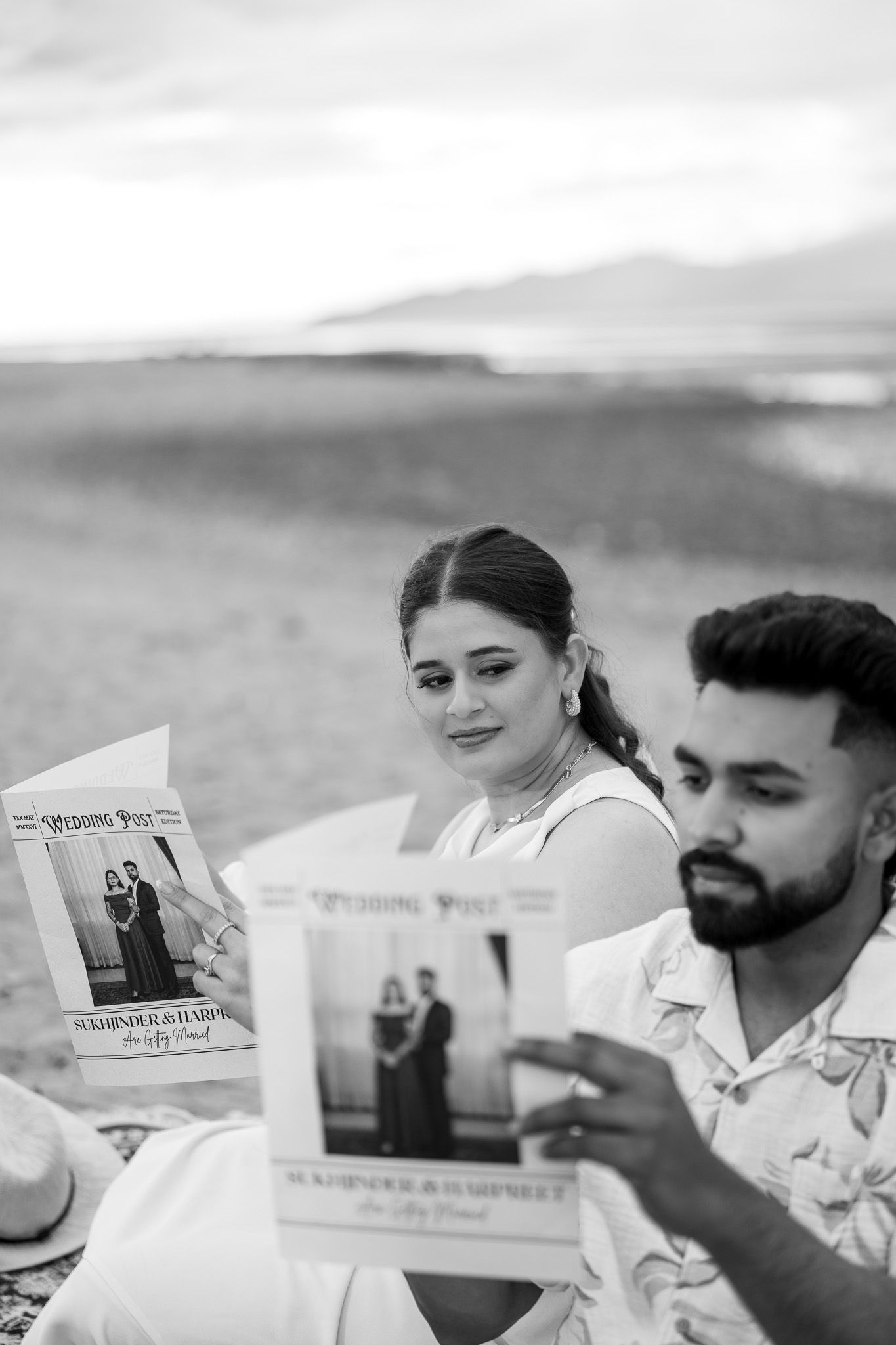 Black and white photo of couple reading Wedding Post newspapers on a beach at sunset.