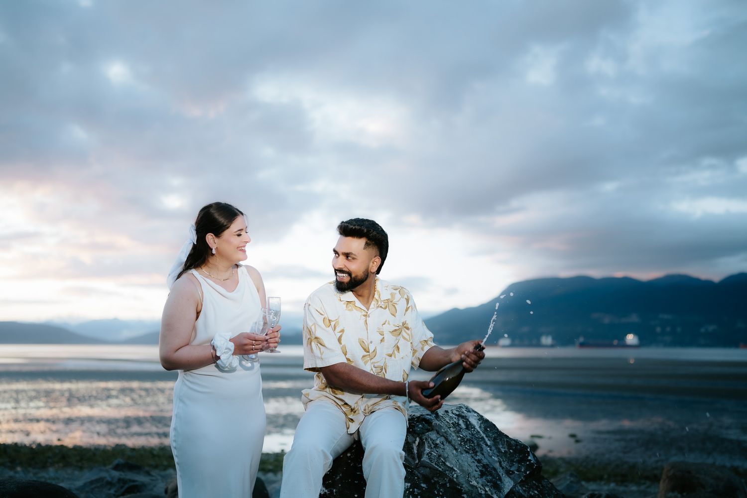 Couple in white and floral outfits standing on rocks by the ocean at sunset with mountains in the background.
