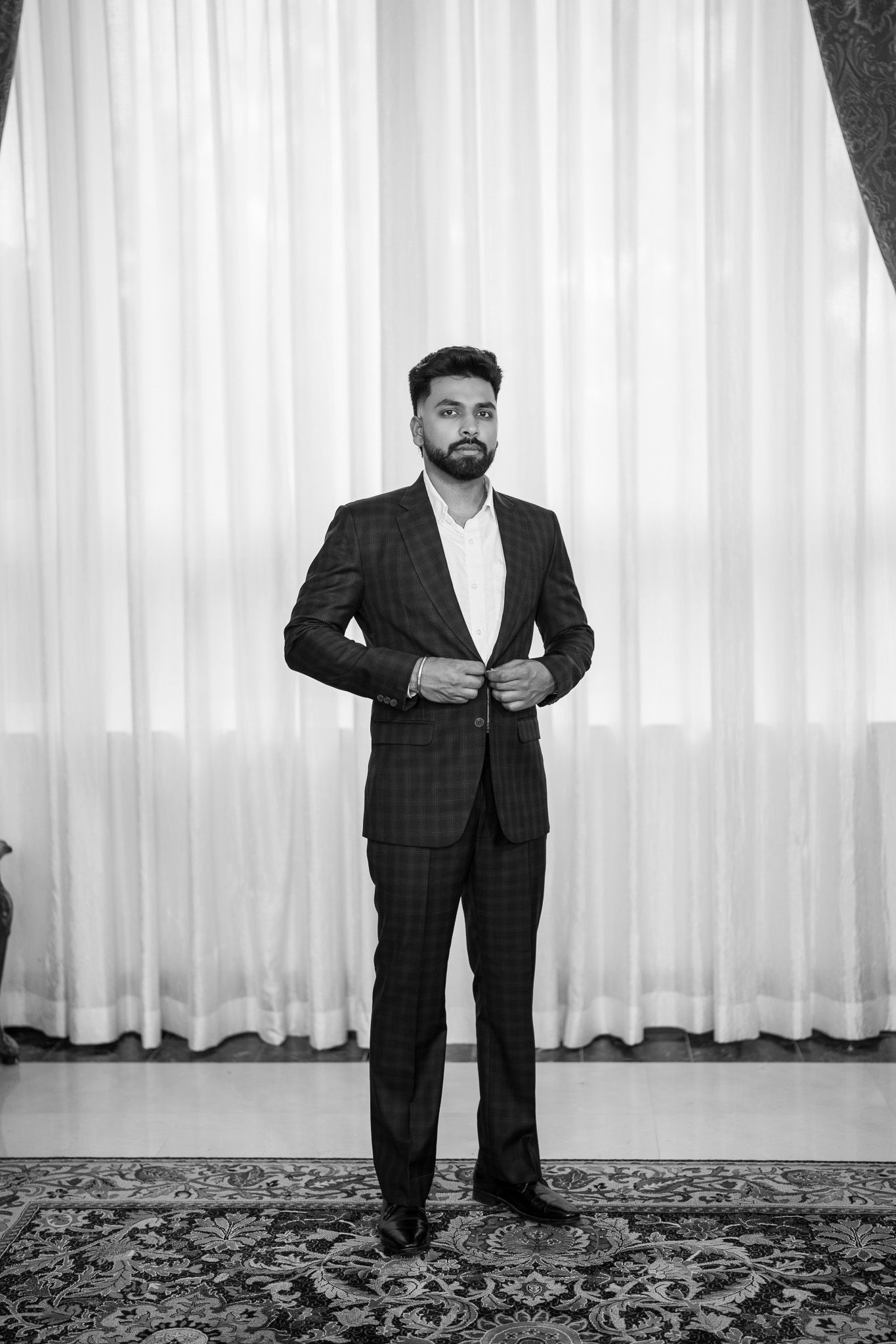 Stylish bearded man in plaid suit buttoning jacket, standing on ornate rug before white curtains, black and white photo.