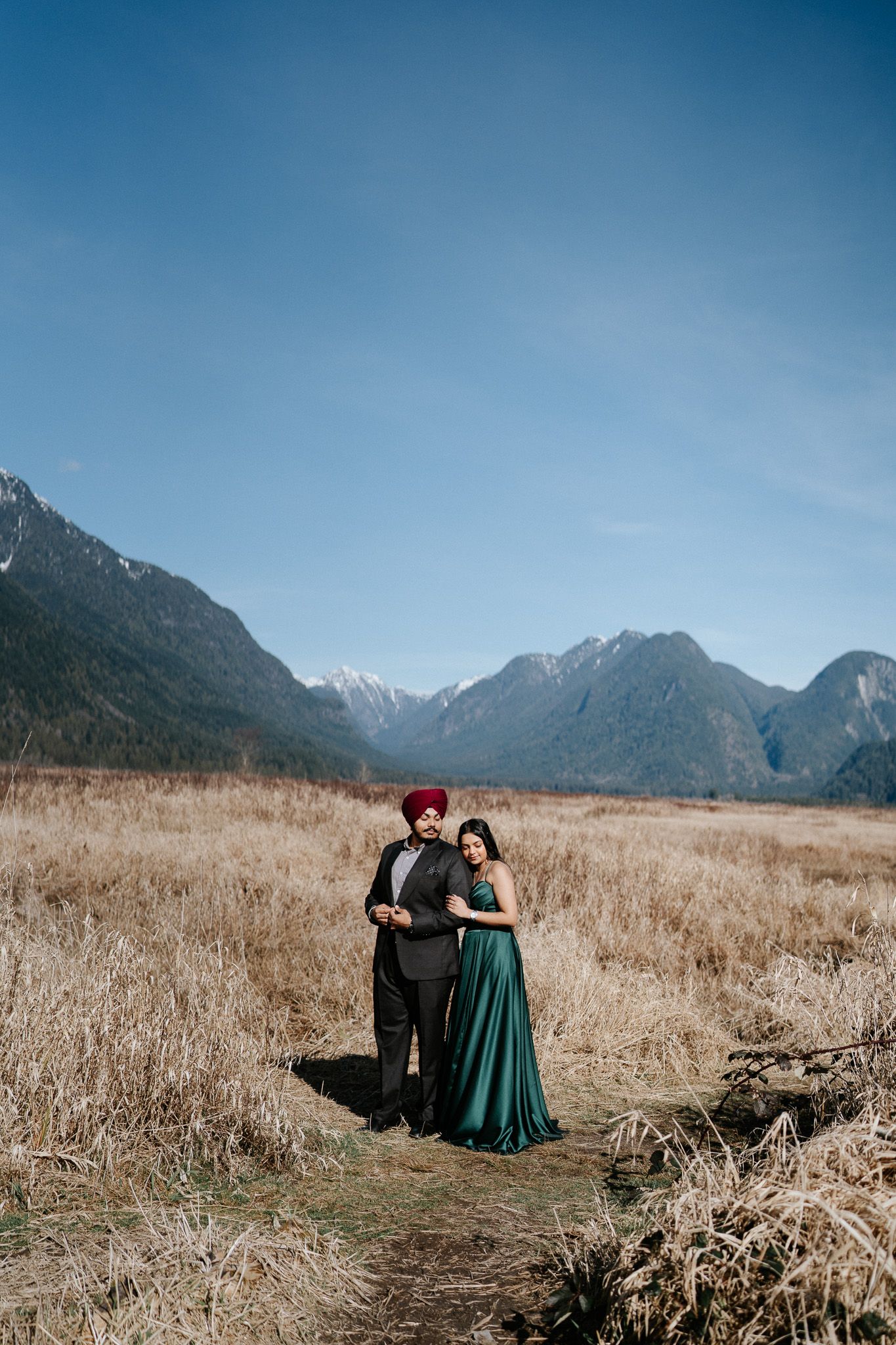 A couple in formal attire embraces in a scenic mountain meadow with dramatic peaks and golden grass in the background.