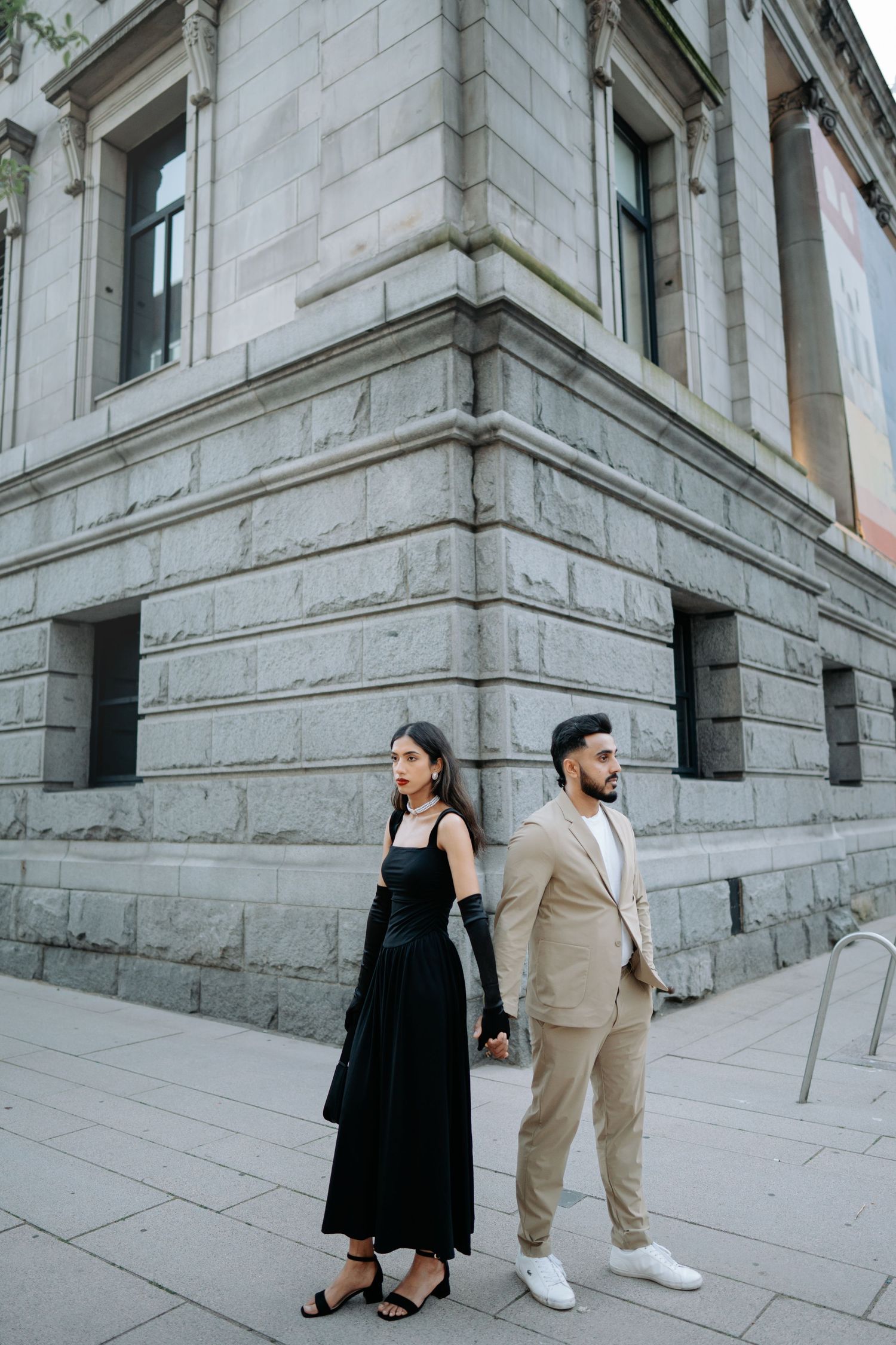 Couple in contrasting black and beige outfits pose on stone steps of a grand building.
