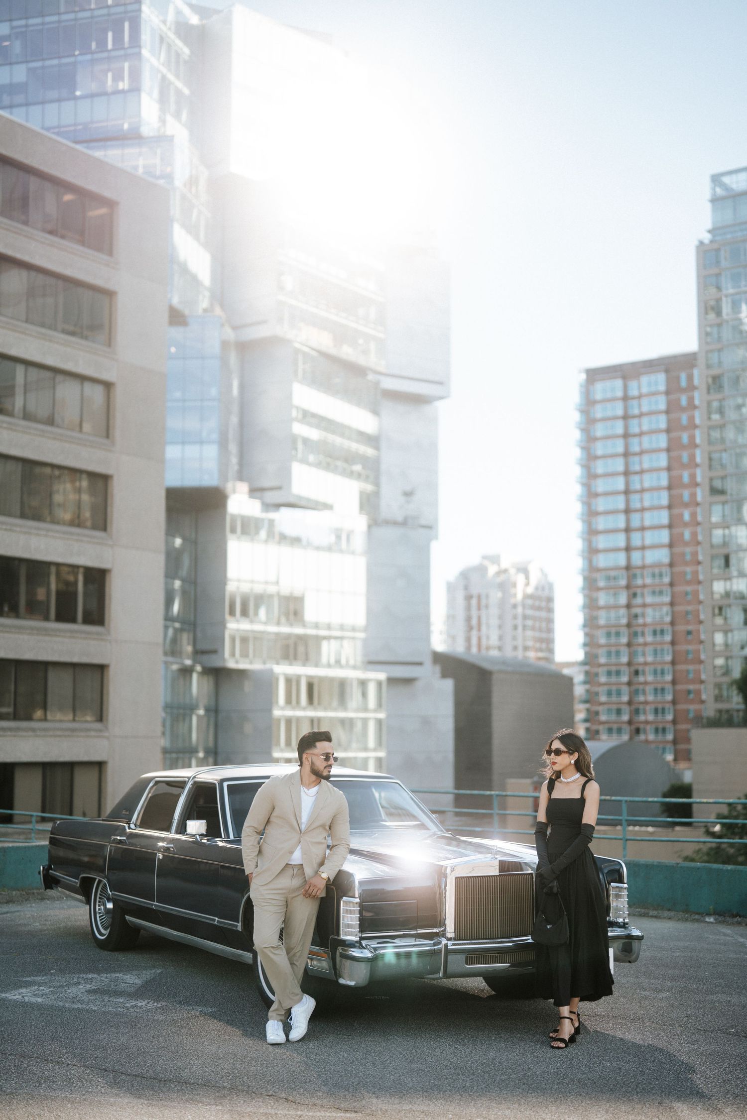 A stylish couple poses near a vintage black limousine on a modern urban rooftop with glass buildings in the background.
