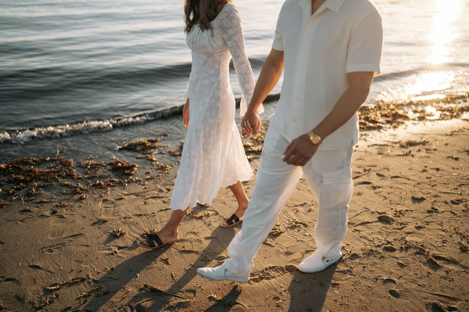 A couple in white outfits holding hands while walking on a sandy beach at sunset by the ocean.