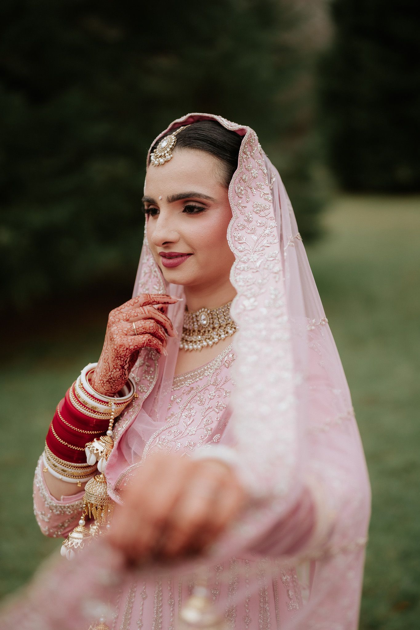 Traditional bride in pink embroidered attire with henna adorned hands poses gracefully in an outdoor setting.