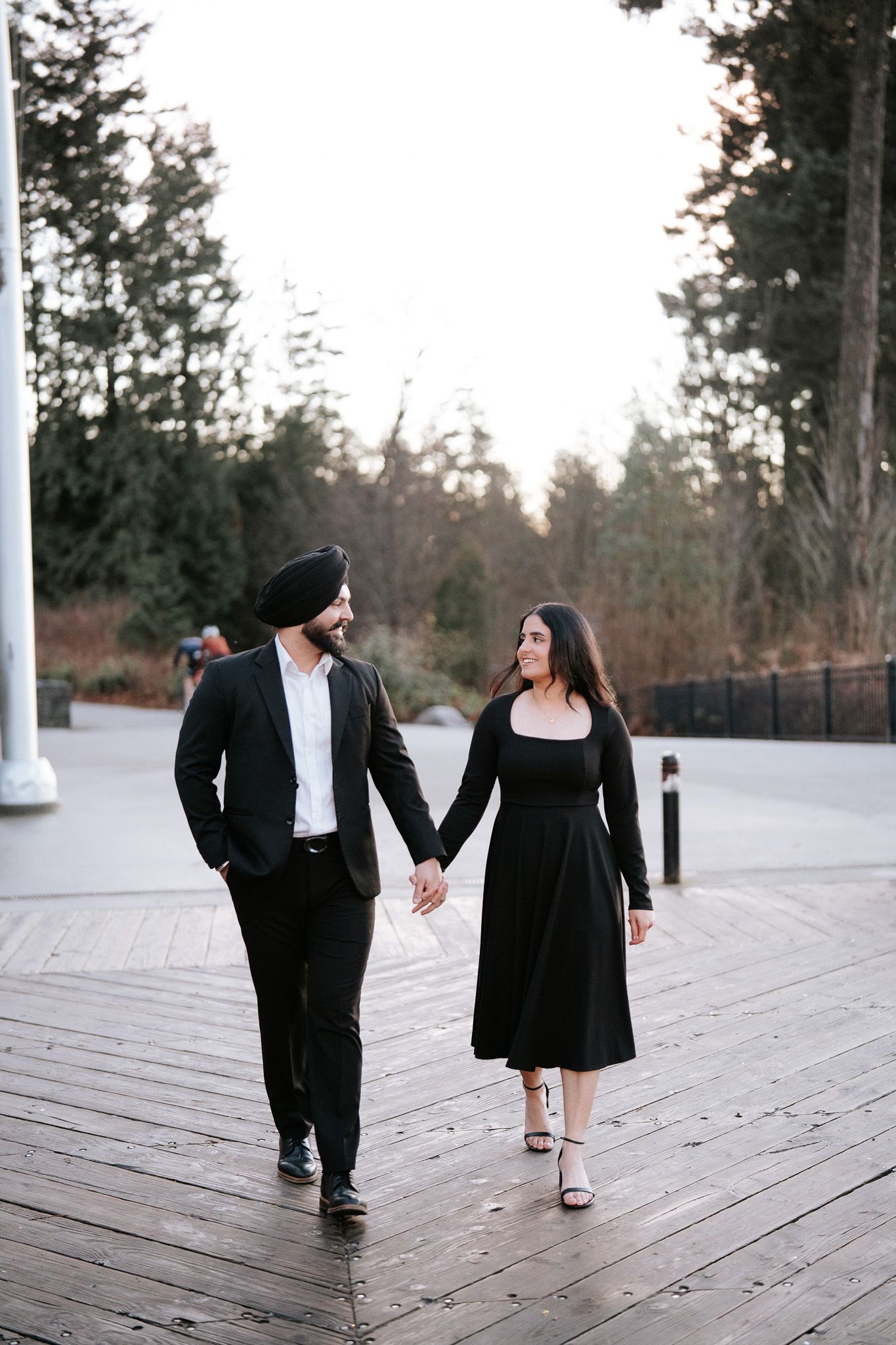 Couple in elegant black attire holding hands while walking together on a wooden boardwalk at dusk.