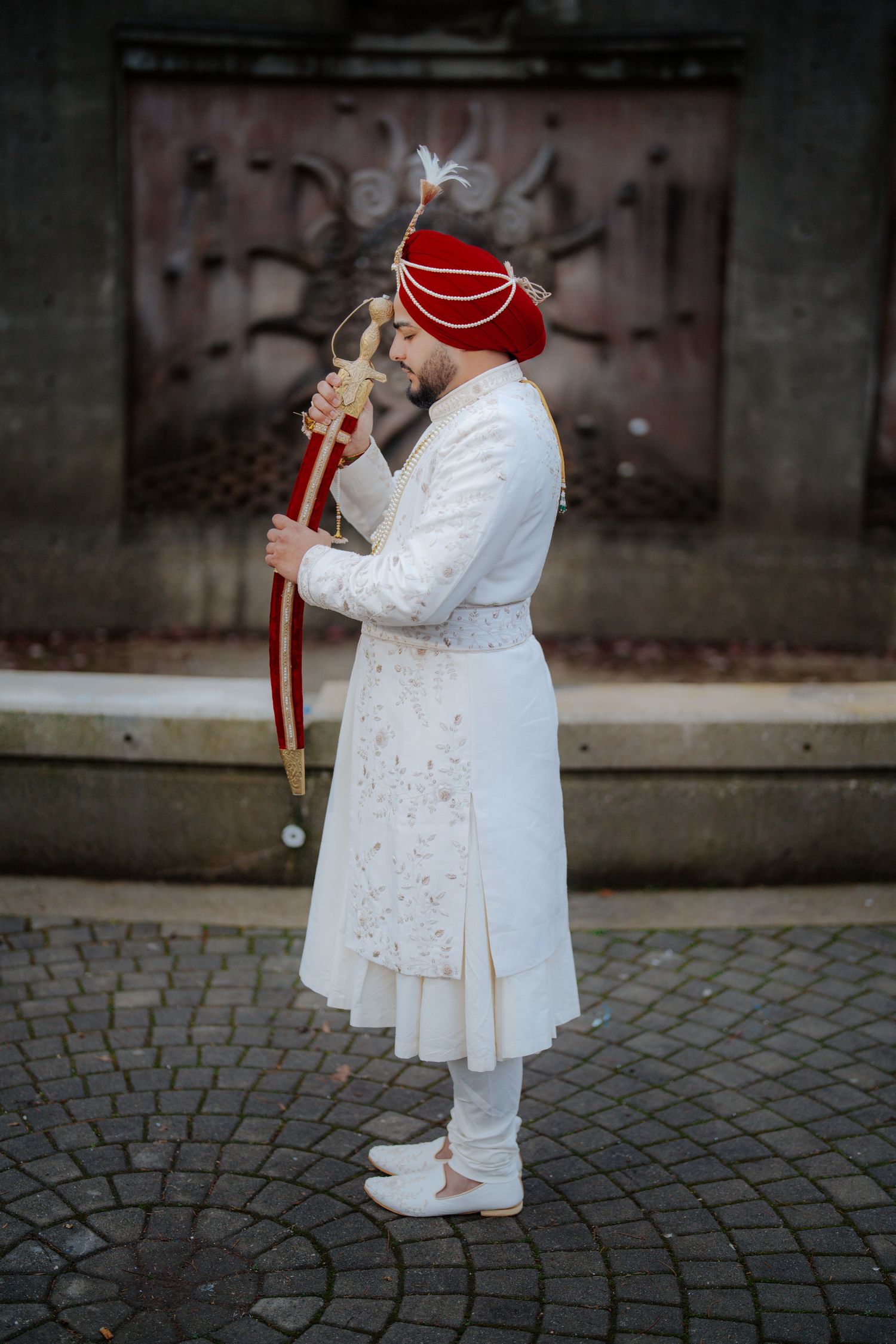 A person in an elegant white outfit and red headpiece poses on cobblestone pavement.
