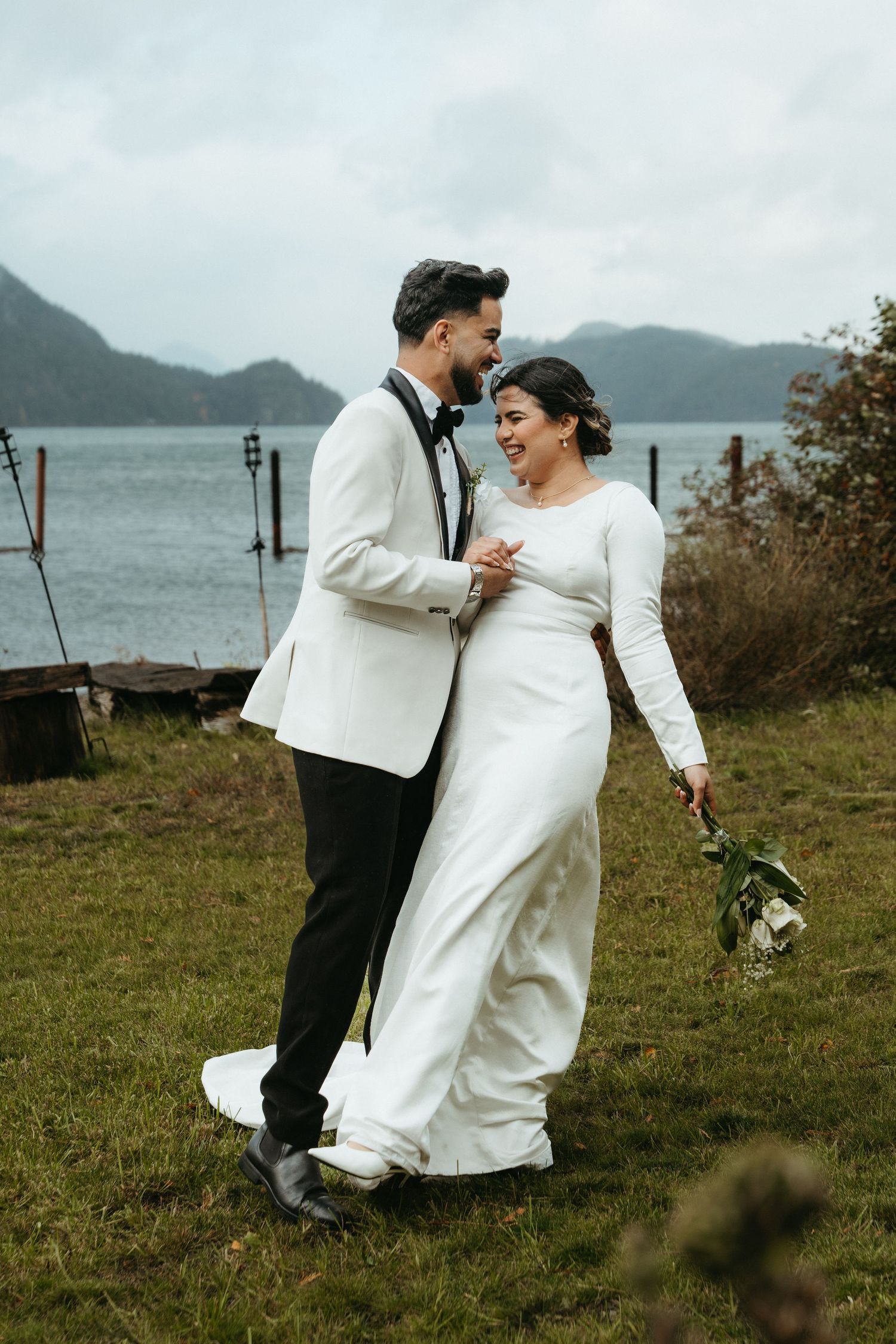 Newlyweds share joyful moments together on a grassy cliff overlooking the ocean during their wedding photoshoot.