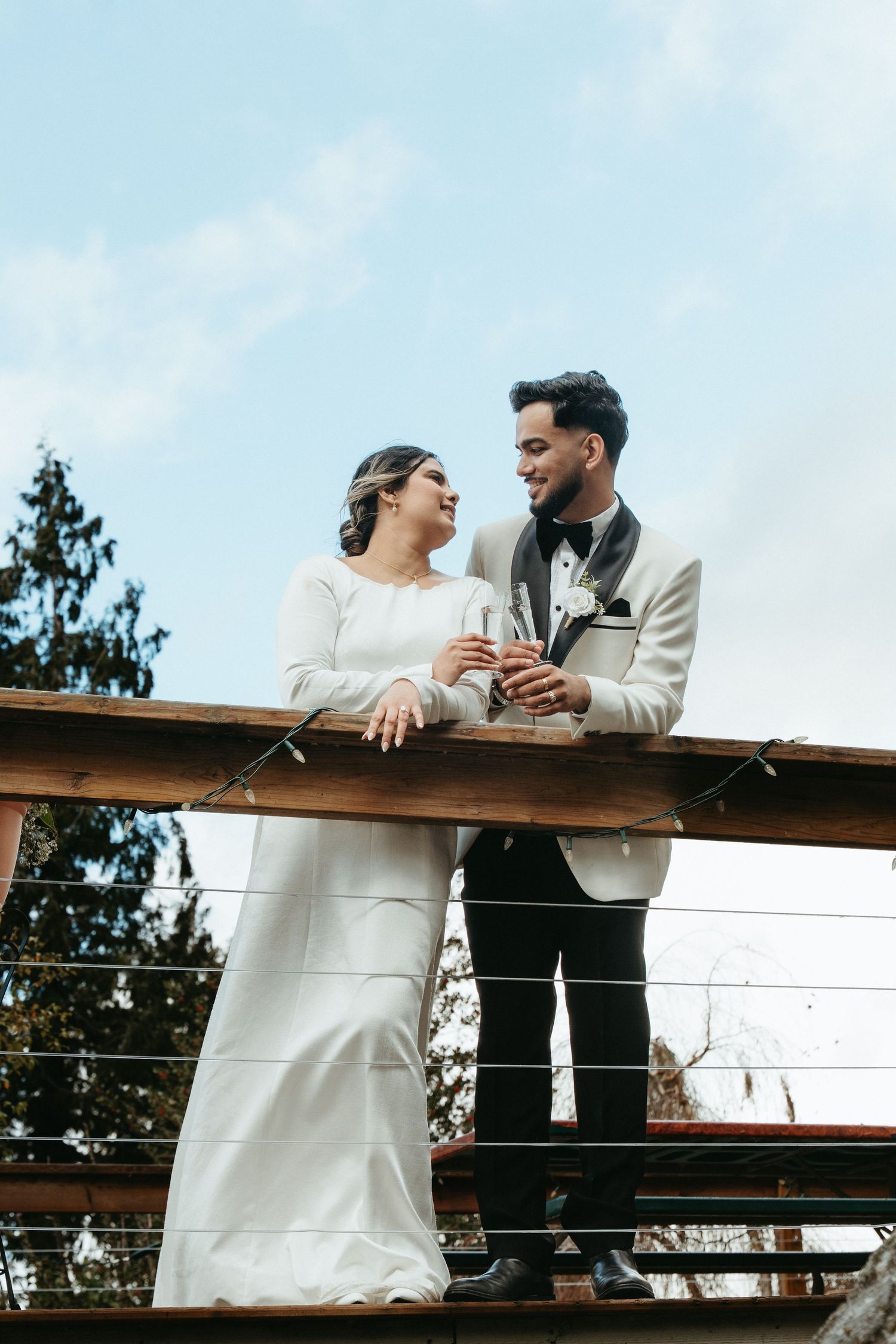 A couple shares a joyful moment on a wooden deck while holding champagne glasses against a blue sky.