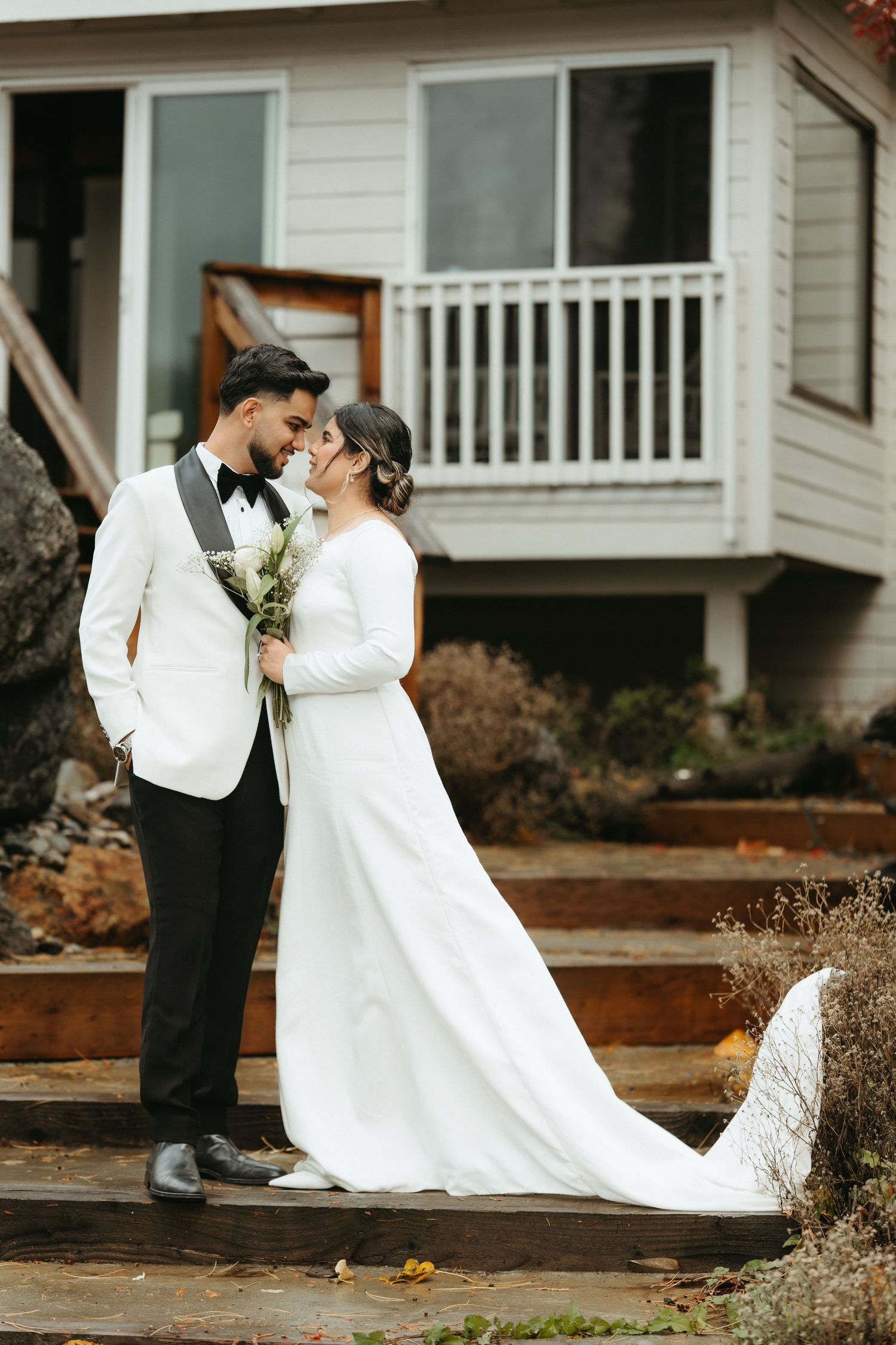 A wedding couple shares an intimate moment outside a white house with a porch in a romantic outdoor setting.