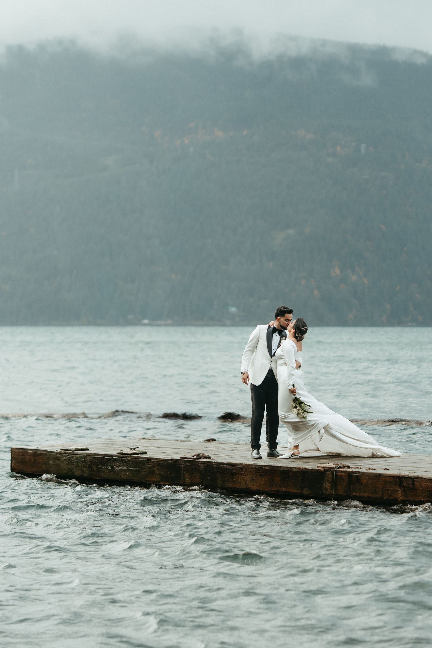 A couple in wedding attire stands together on a wooden dock overlooking misty mountains and a serene lake.