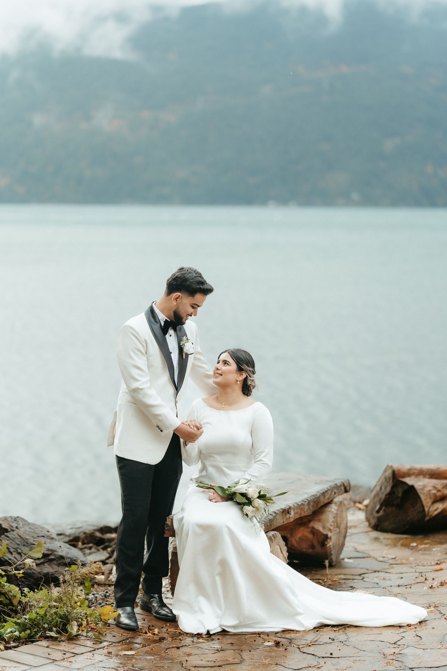 A romantic wedding couple sits on rocky shoreline by misty lake in elegant white dress and dark suit.