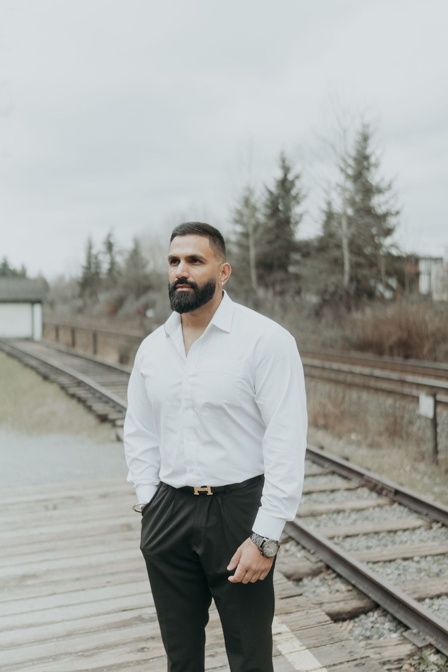 Man in formal attire stands confidently by railway tracks with evergreen trees in background.